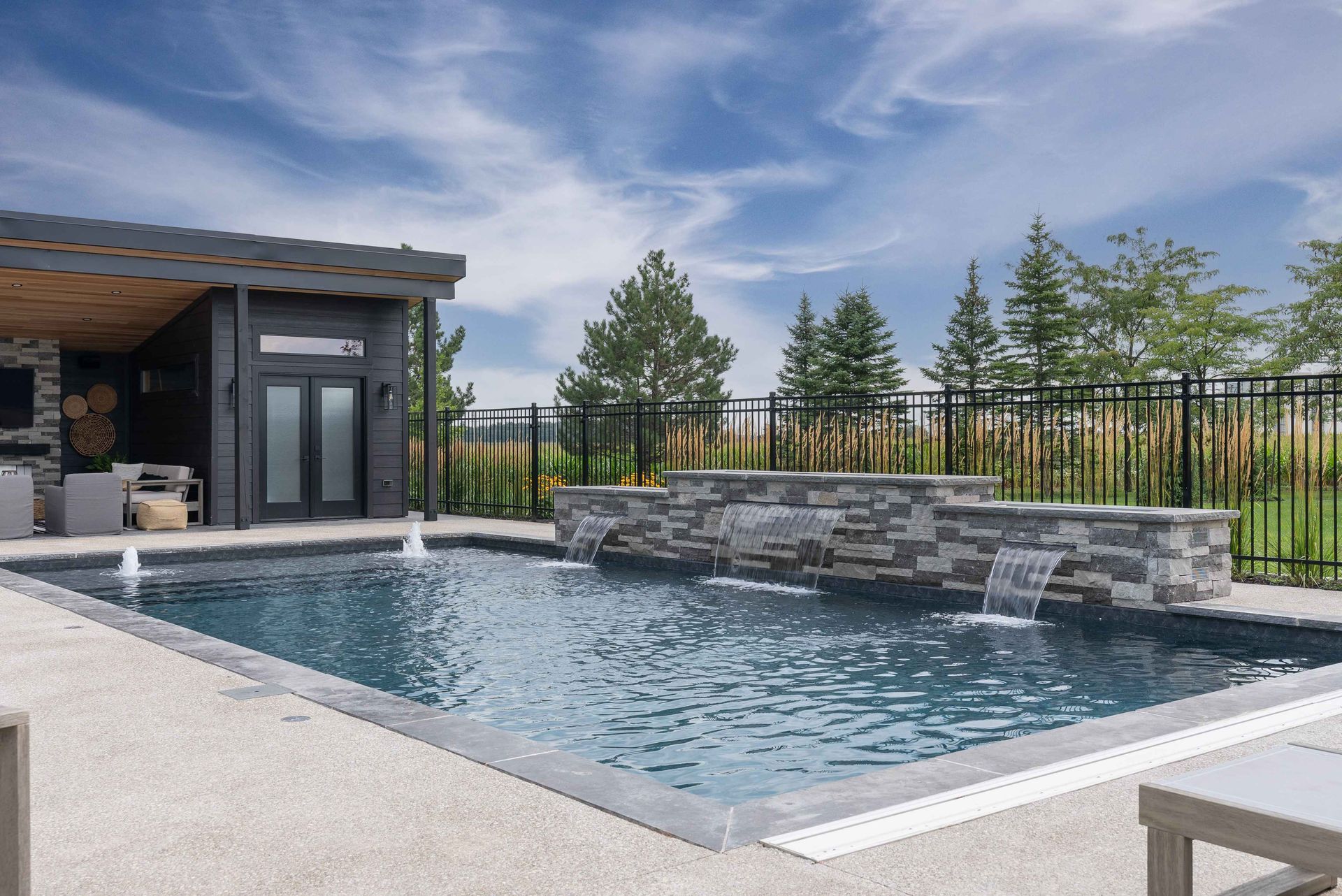 Modern backyard pool with stone water feature, black fence, and patio beside a house under a cloudy sky
