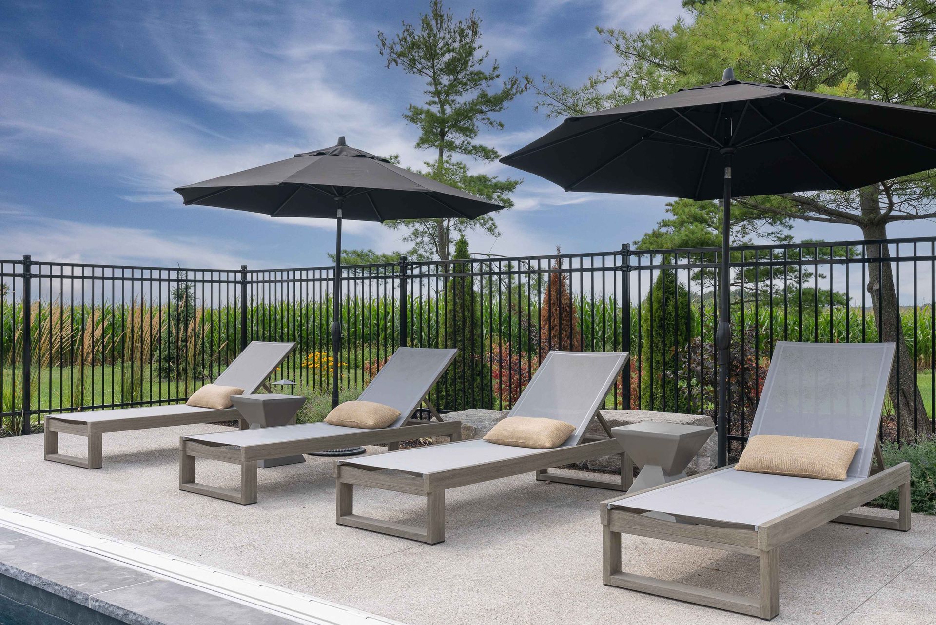 Poolside lounge chairs with gray cushions and black umbrellas beside a fenced patio