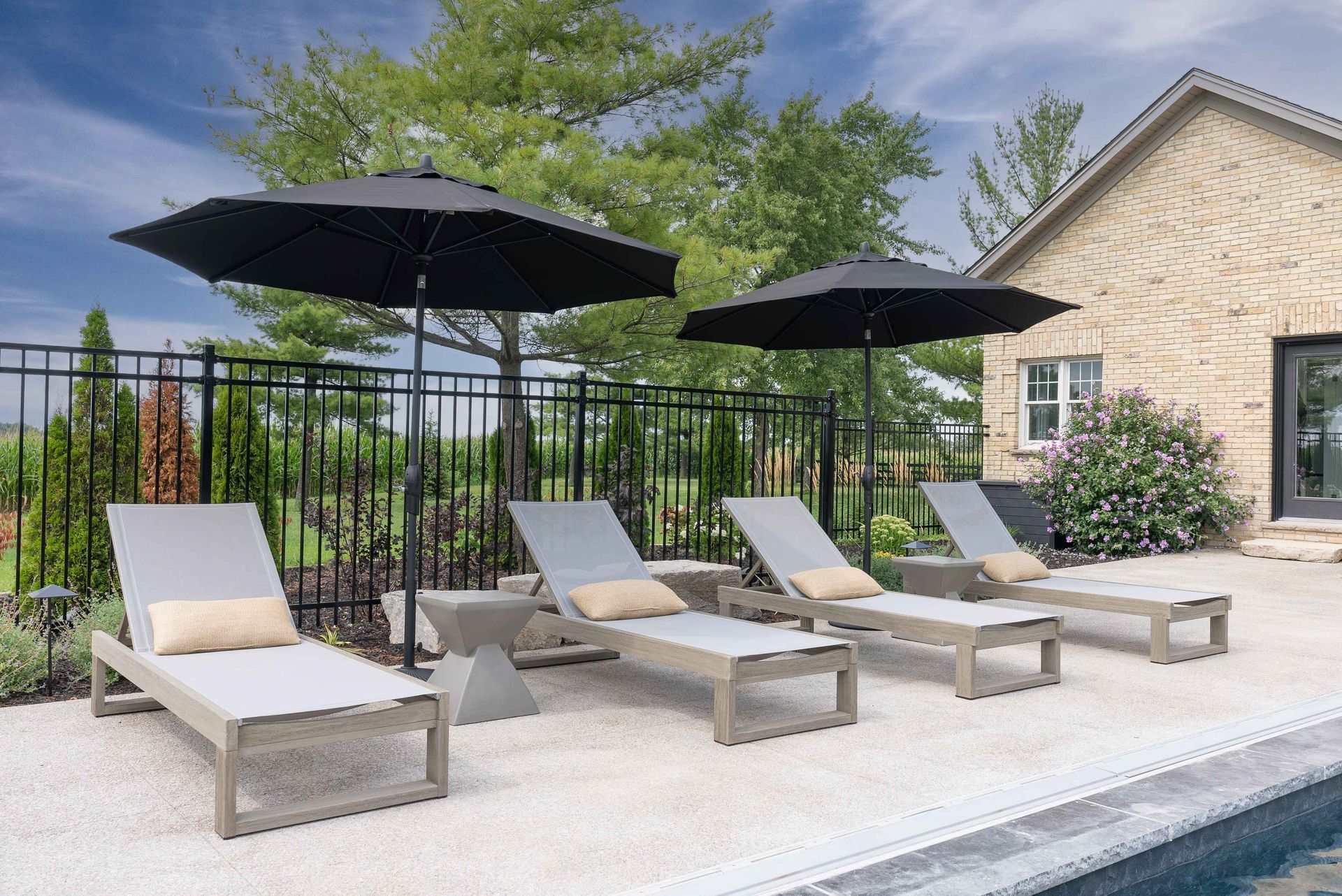 Poolside lounge chairs with black umbrellas beside a stone house and fence