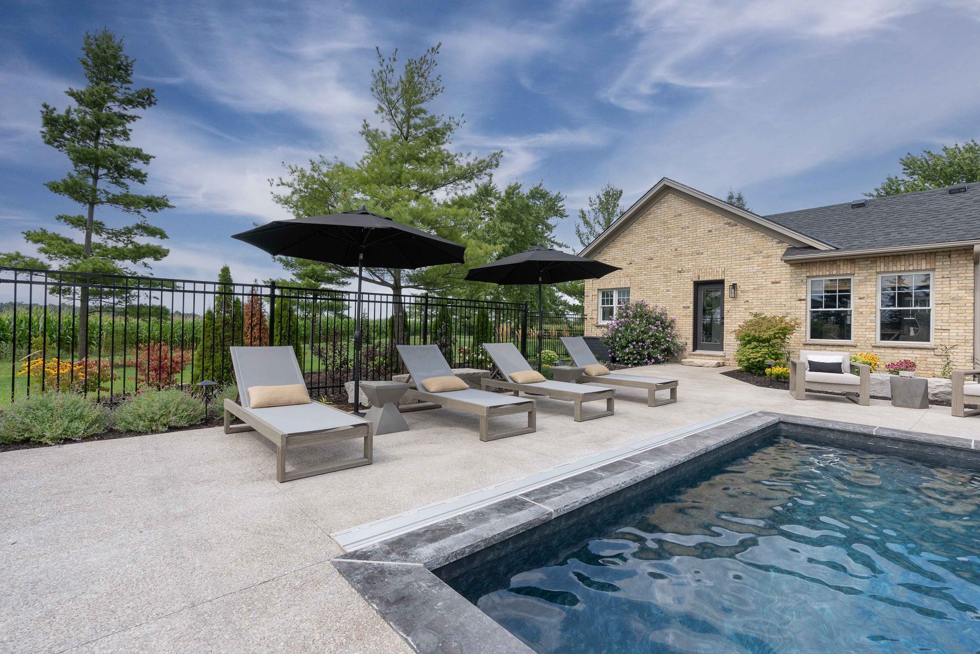 Backyard pool patio with lounge chairs, umbrellas, and a stone house under a blue sky