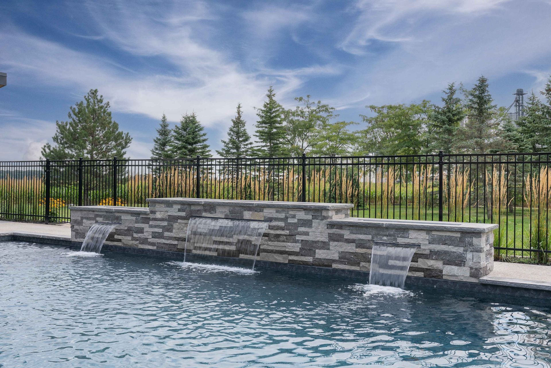 Backyard pool with stone water feature, black fence, and trees under a cloudy sky