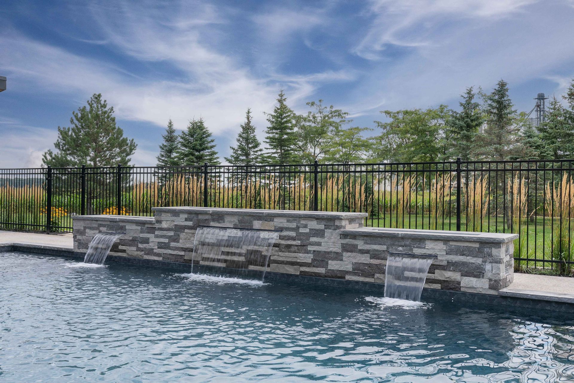 Stone water feature beside a pool with black fence, trees, and blue sky