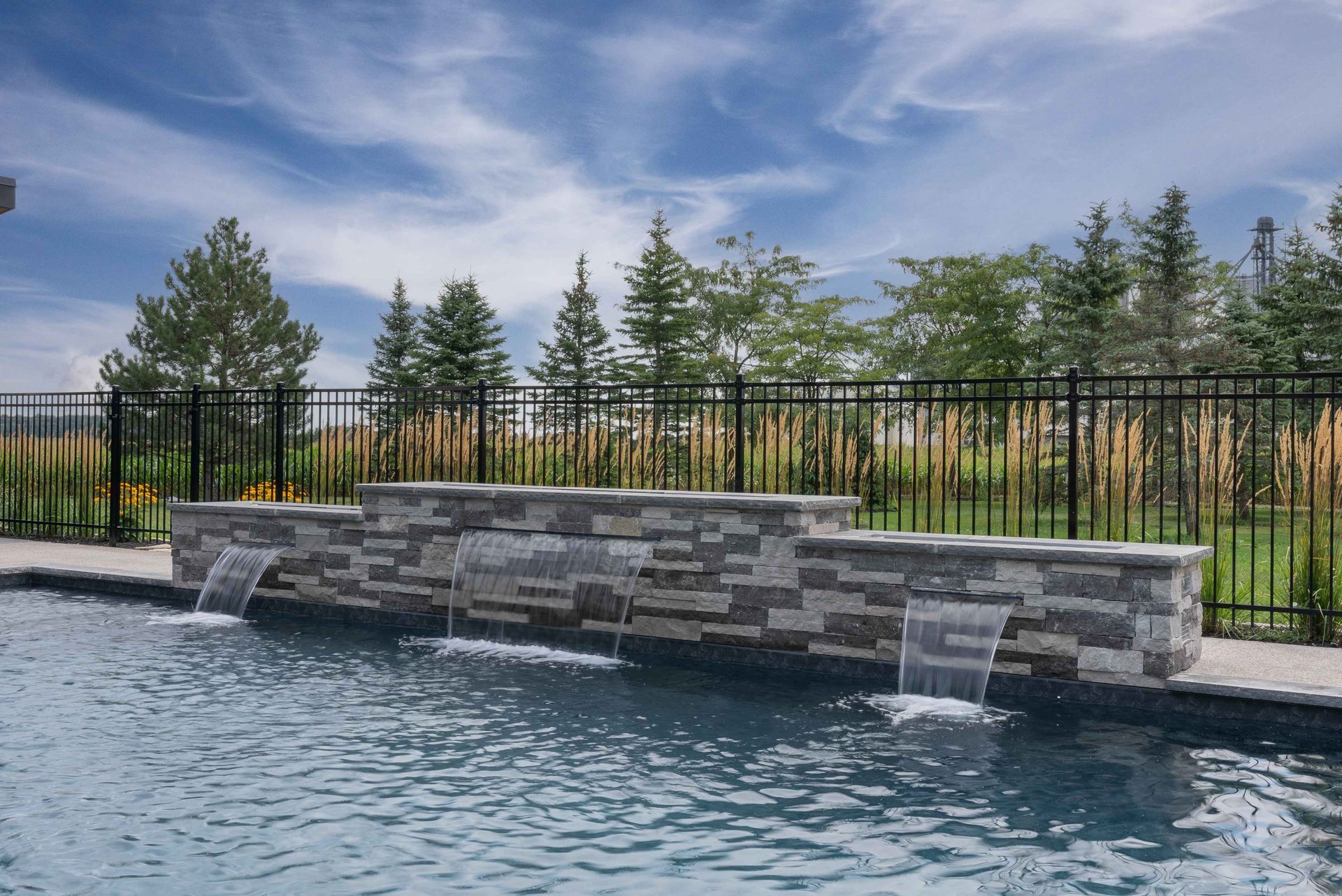 Stone waterfall feature beside a pool, with black fence and trees under a cloudy sky