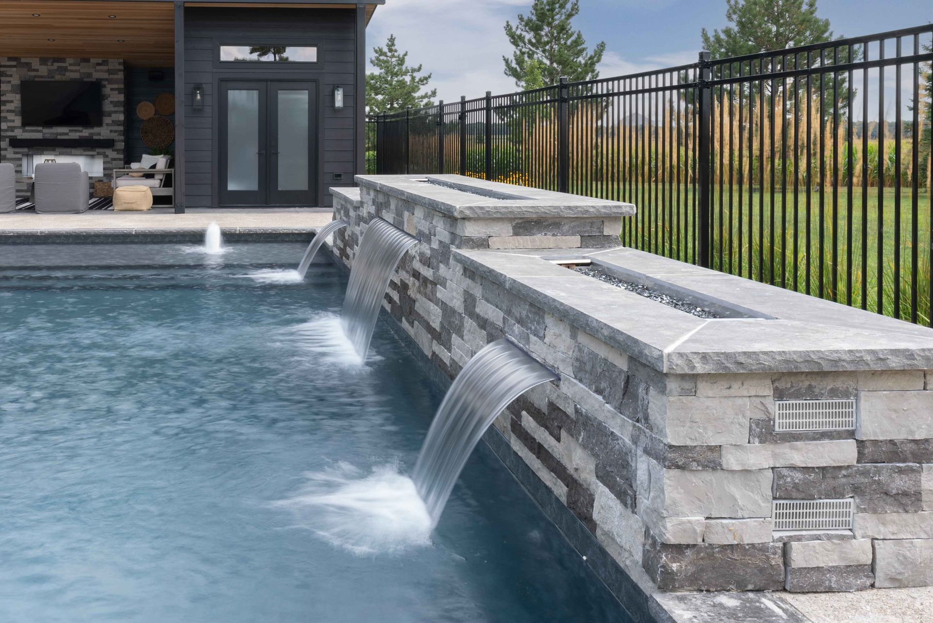 Modern pool with stone water features and a black metal fence beside a patio and house