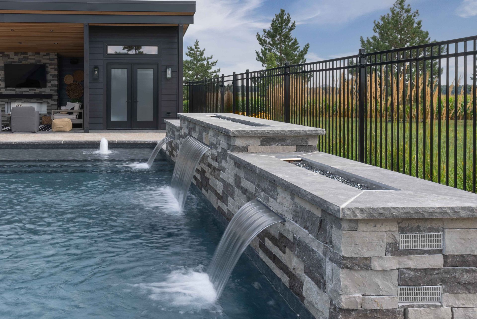 Modern backyard pool with stone waterfall wall, black fence, and a patio building in the background