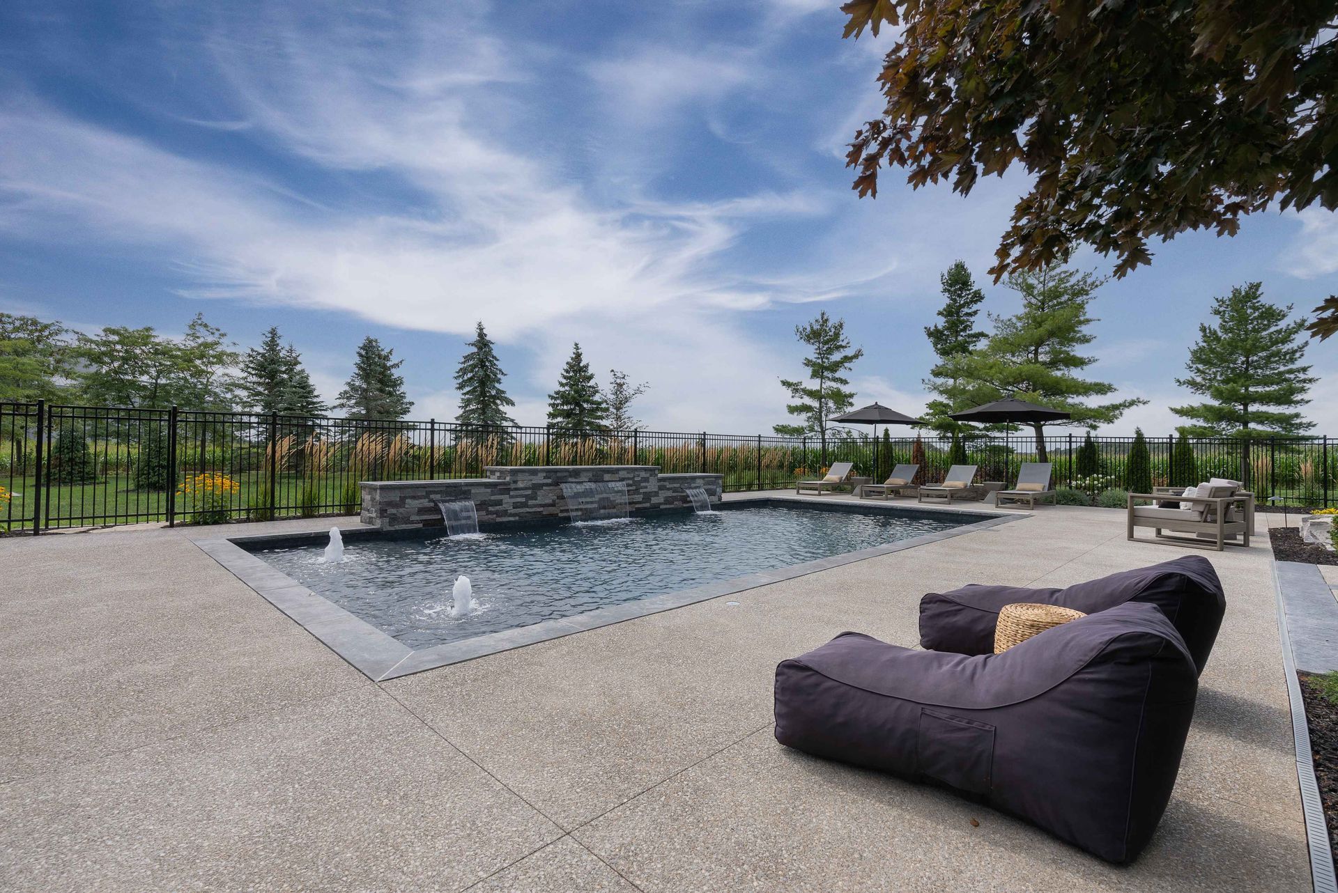 Modern backyard patio with a pool, black lounge chairs, and trees under a blue sky