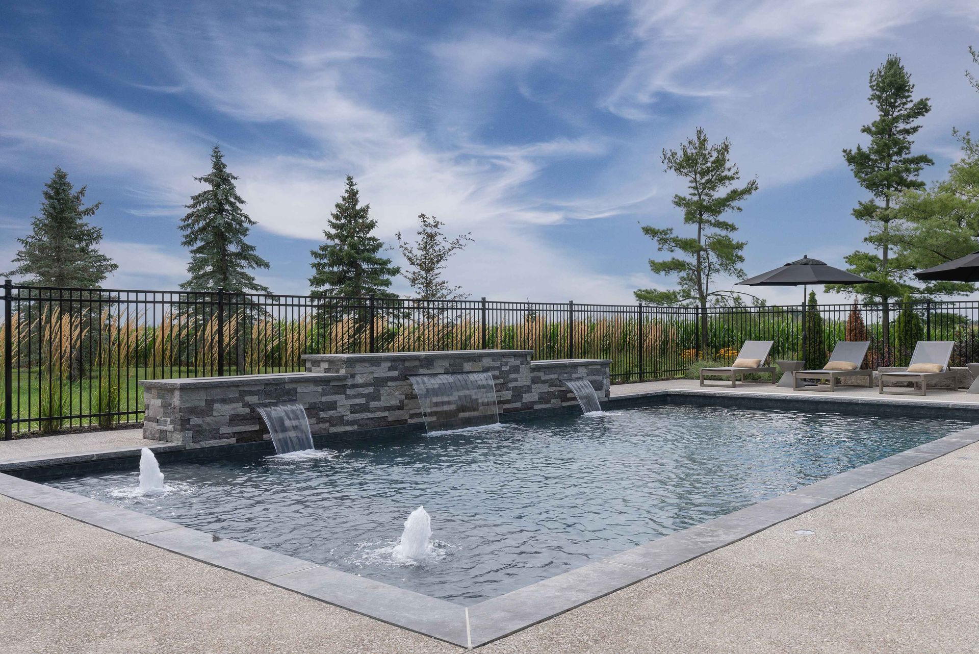Outdoor pool with stone waterfall, white fountains, lounge chairs, and trees under a blue sky
