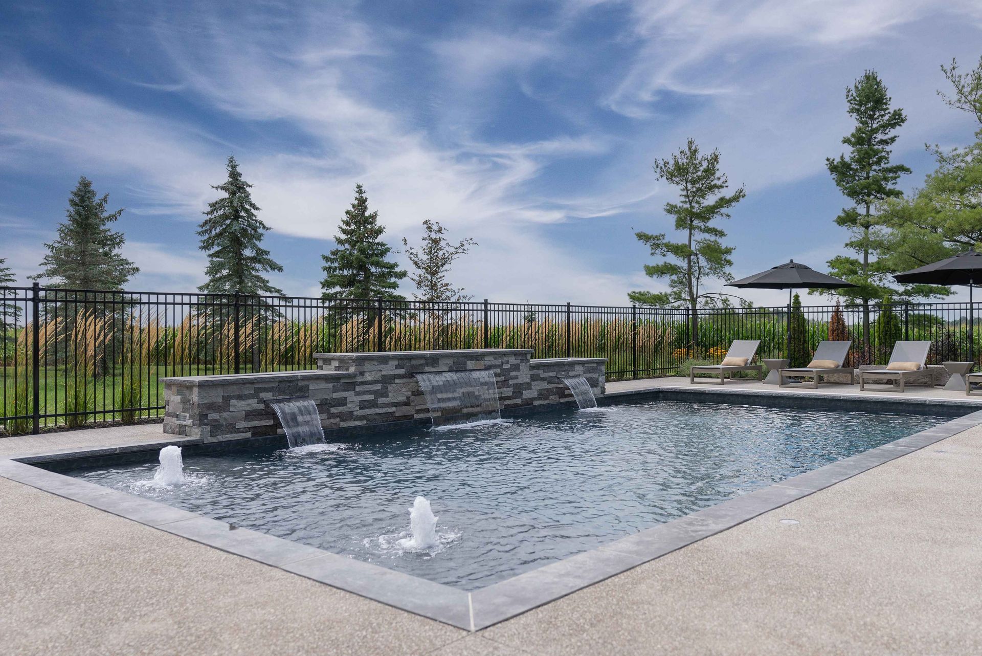 Modern outdoor pool with stone fountain, lounge chairs, and fenced patio under a blue sky