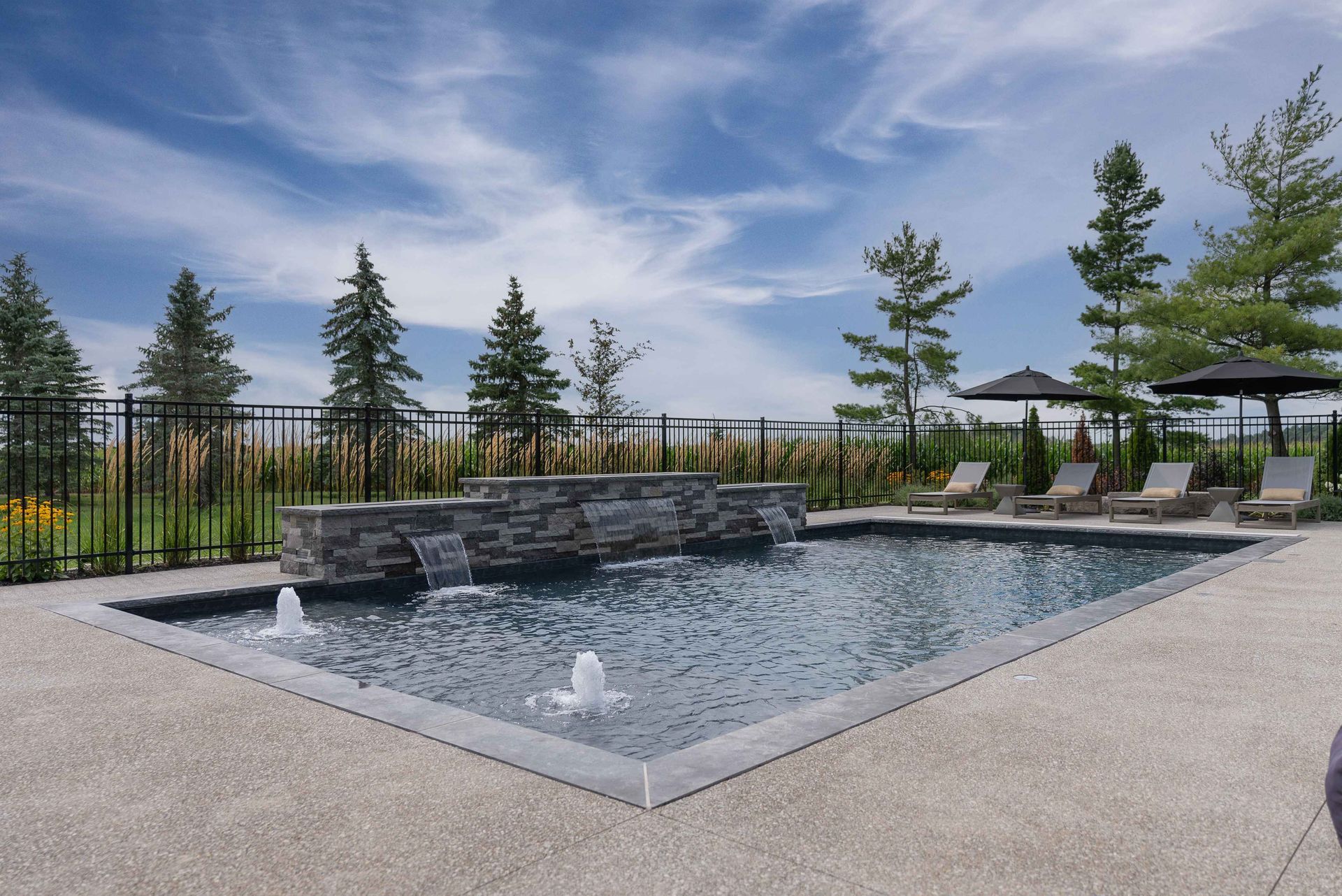 Modern outdoor pool with stone waterfall, lounge chairs, and trees under a cloudy sky