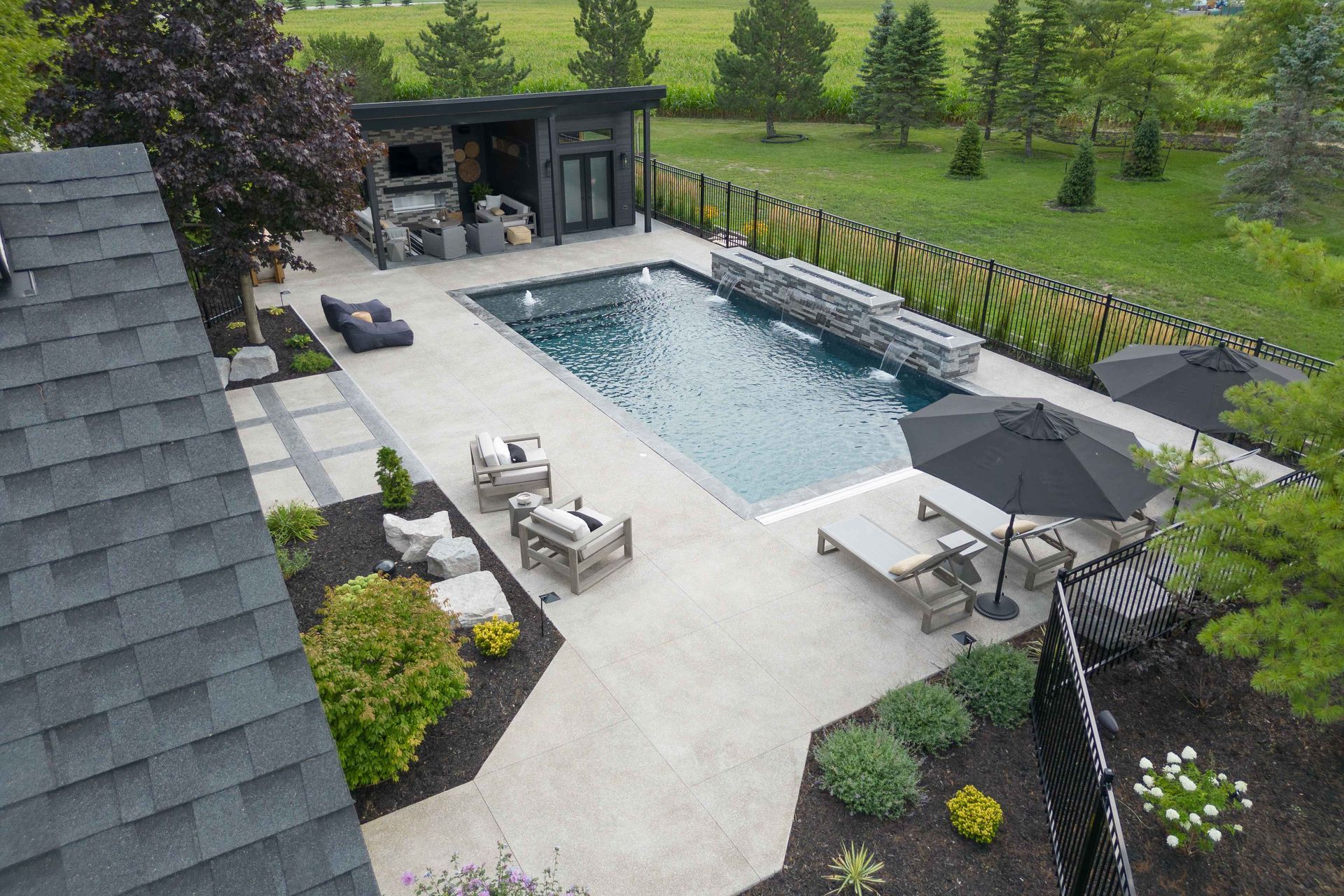 Modern backyard pool with patio furniture, lounge chairs, umbrellas, and landscaped greenery