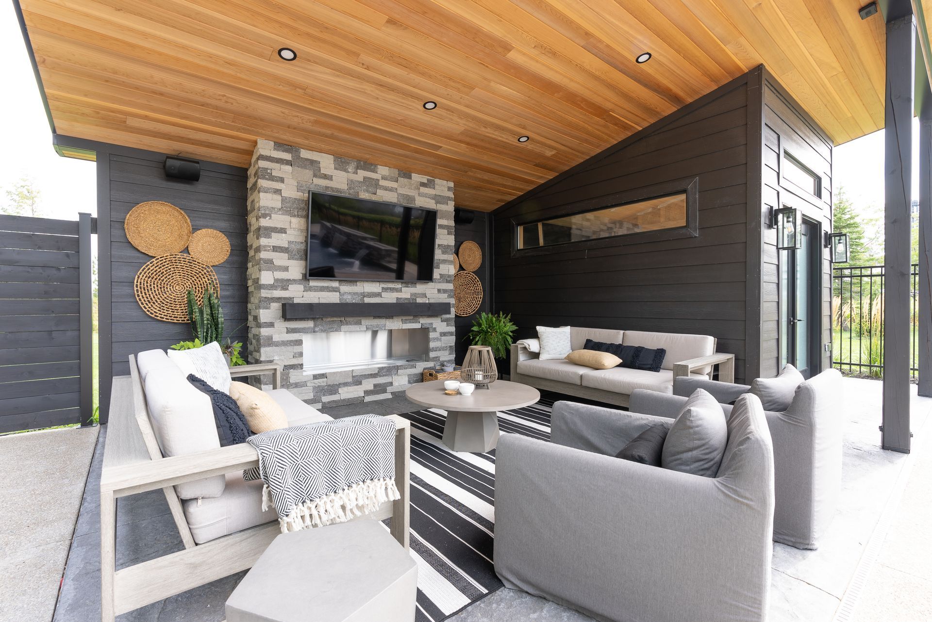 Modern covered patio lounge with gray seating, stone fireplace, striped rug, and wood ceiling