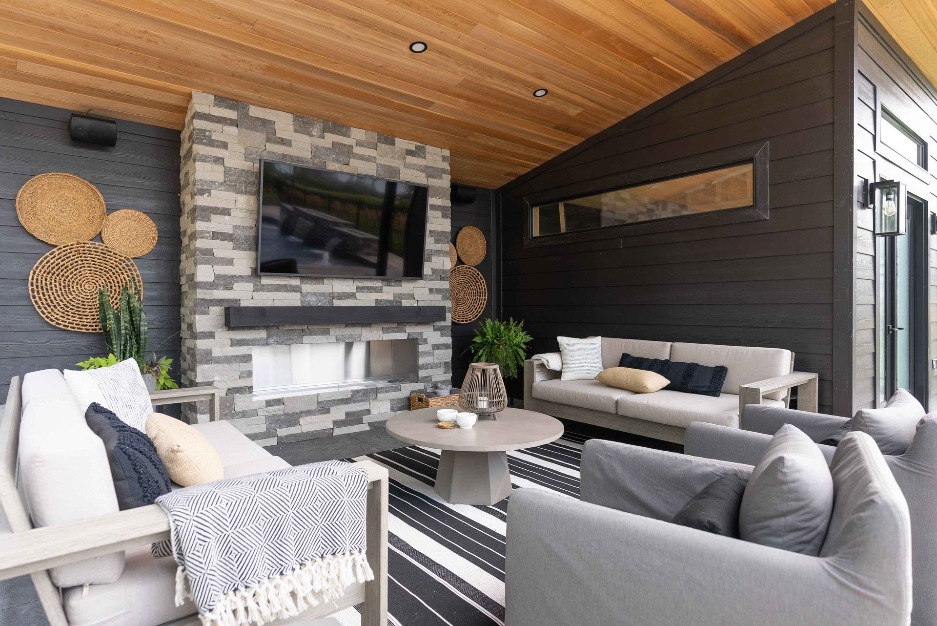 Modern covered patio with gray sofas, stone fireplace, TV, and striped outdoor rug under a wood ceiling