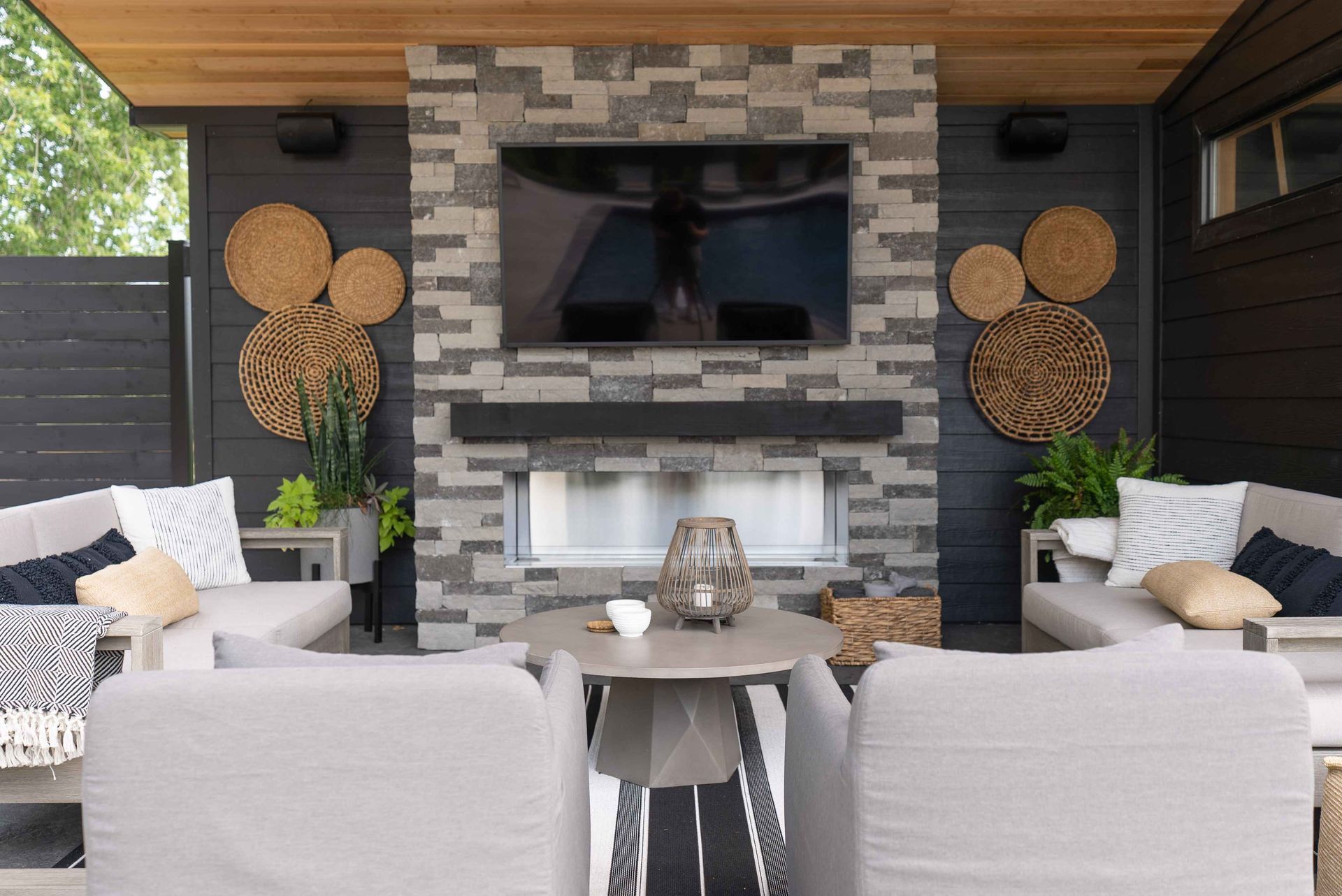 Covered patio seating area with TV and fireplace, gray furniture, wood ceiling, and decorative wall accents