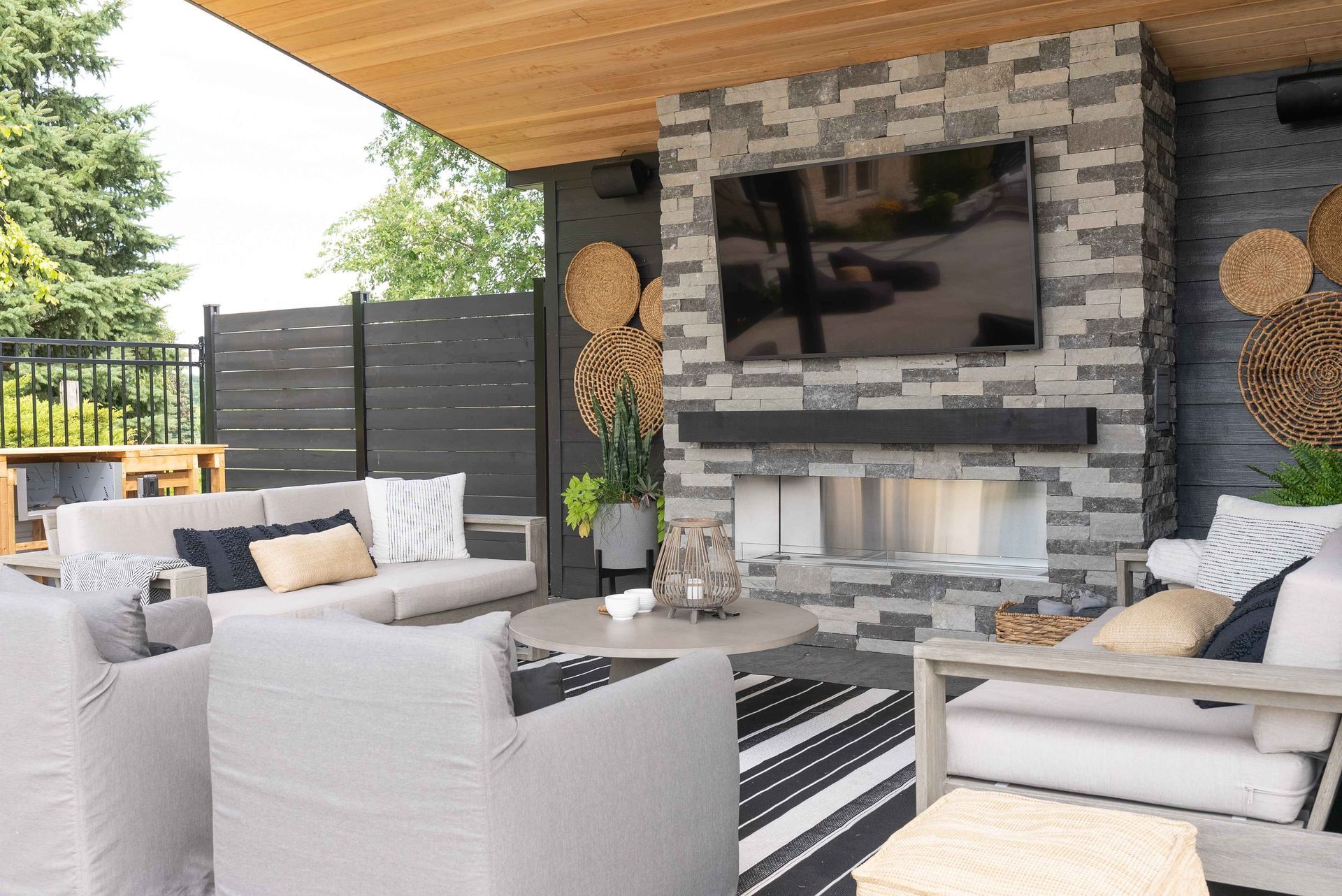 Covered patio lounge with gray sofas, striped rug, and wall-mounted TV over a stone fireplace