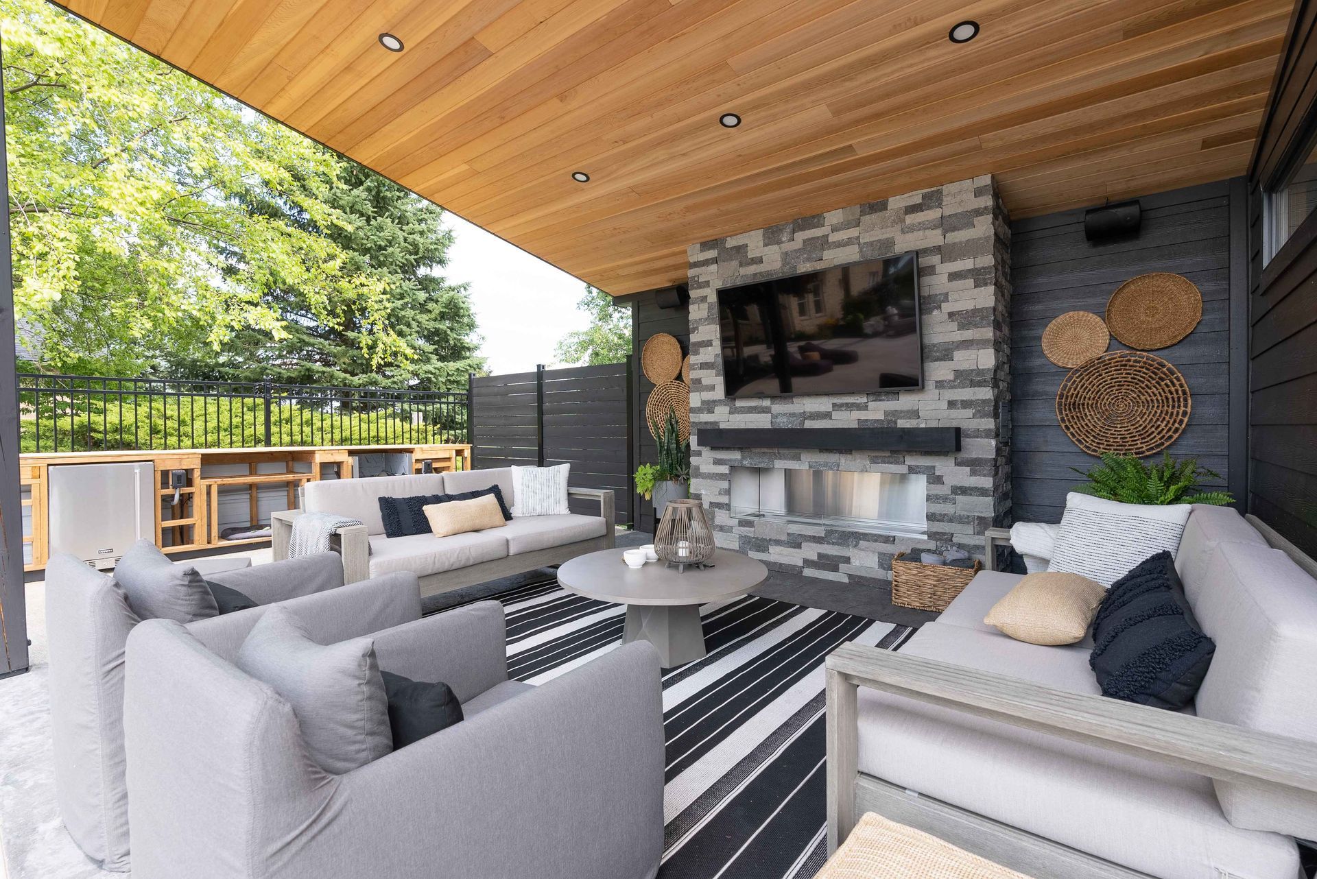 Modern covered patio with gray sofas, striped rug, stone fireplace, and wood ceiling overlooking a green yard