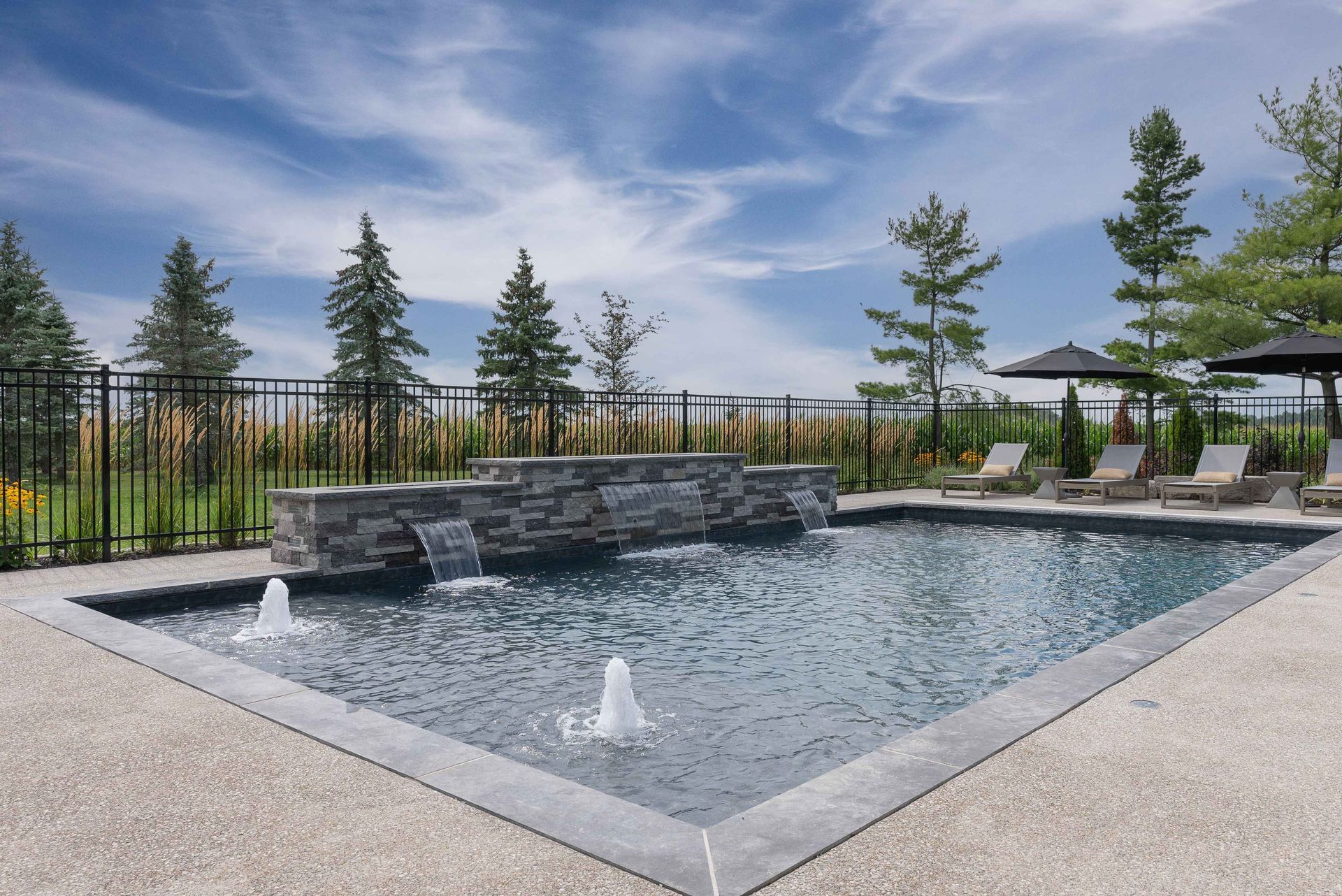 Modern outdoor pool with stone waterfall, fountains, lounge chairs, and trees under a blue sky
