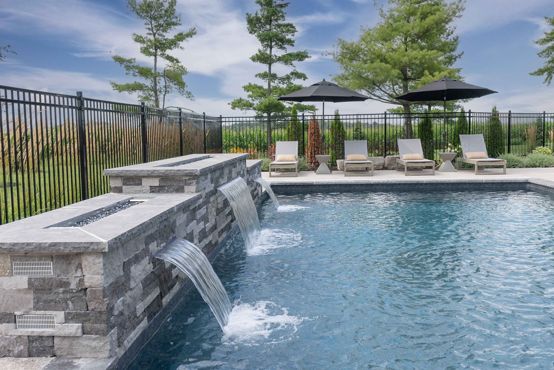 Backyard pool with stone waterfall, lounge chairs, and umbrella seating beside a grassy view