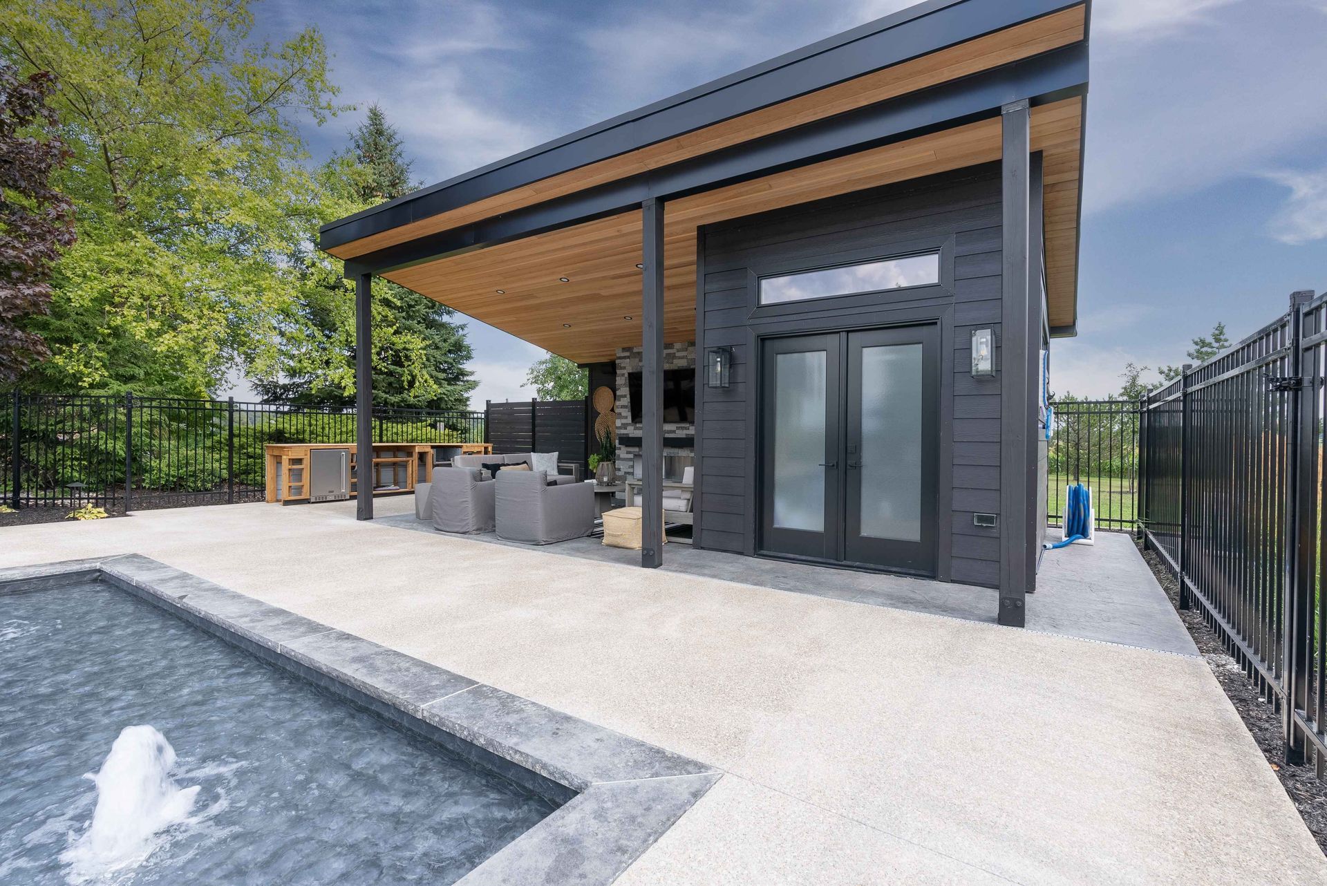 Modern backyard pool patio with a covered outdoor kitchen and glass doors beside a fence