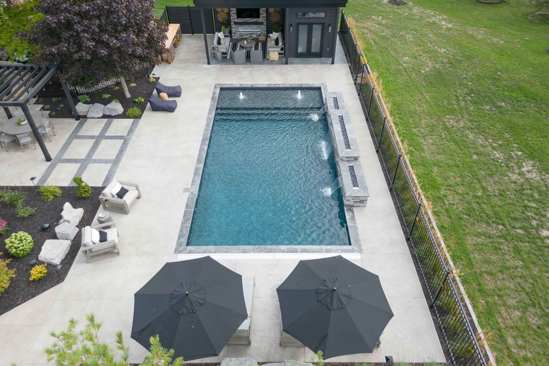 Aerial view of a backyard pool with lounge chairs, umbrellas, and patio seating beside a lawn