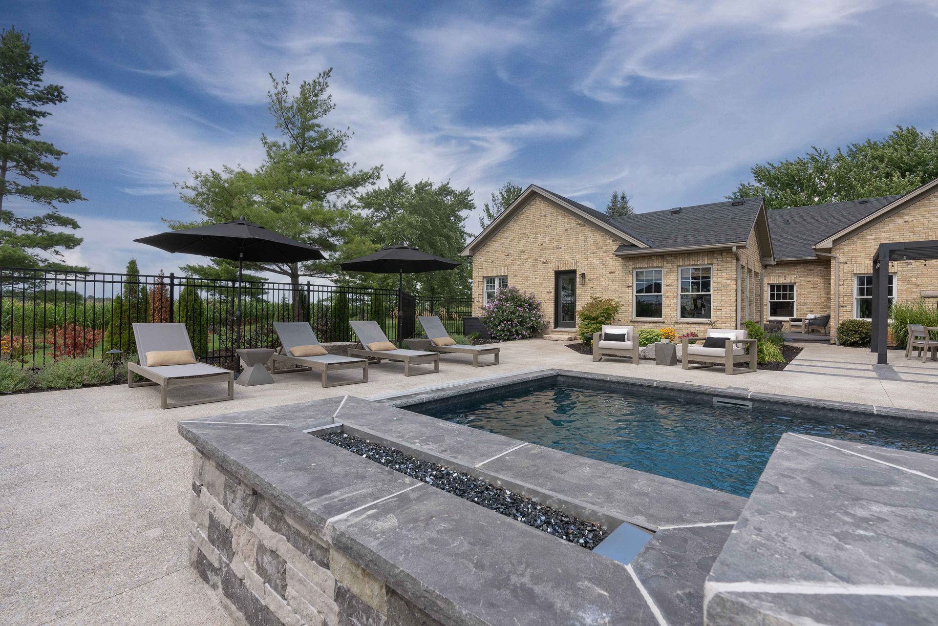 Stone patio with a pool, lounge chairs, and umbrellas beside a beige house under a partly cloudy sky