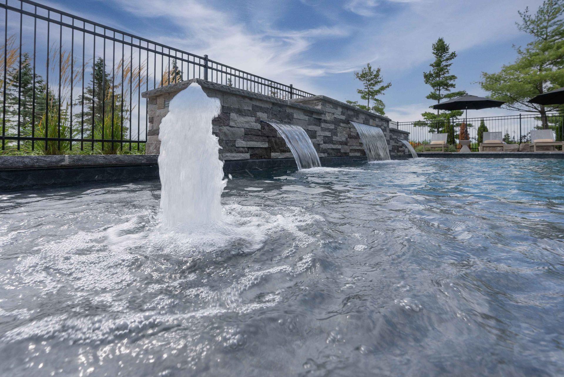 Modern outdoor pool with fountain jets, stone wall, and blue water under a partly cloudy sky