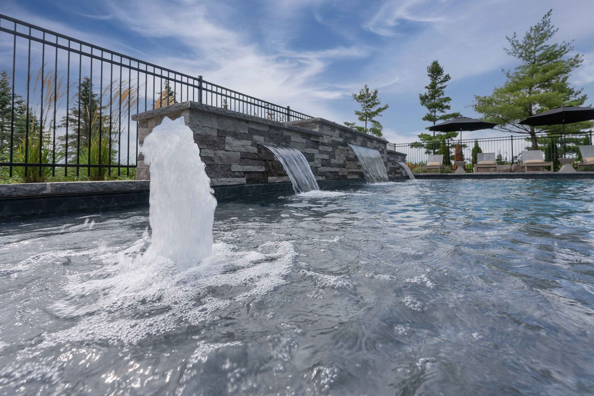 A fountain splashing in a shallow pool beside a stone waterfall under a blue sky.