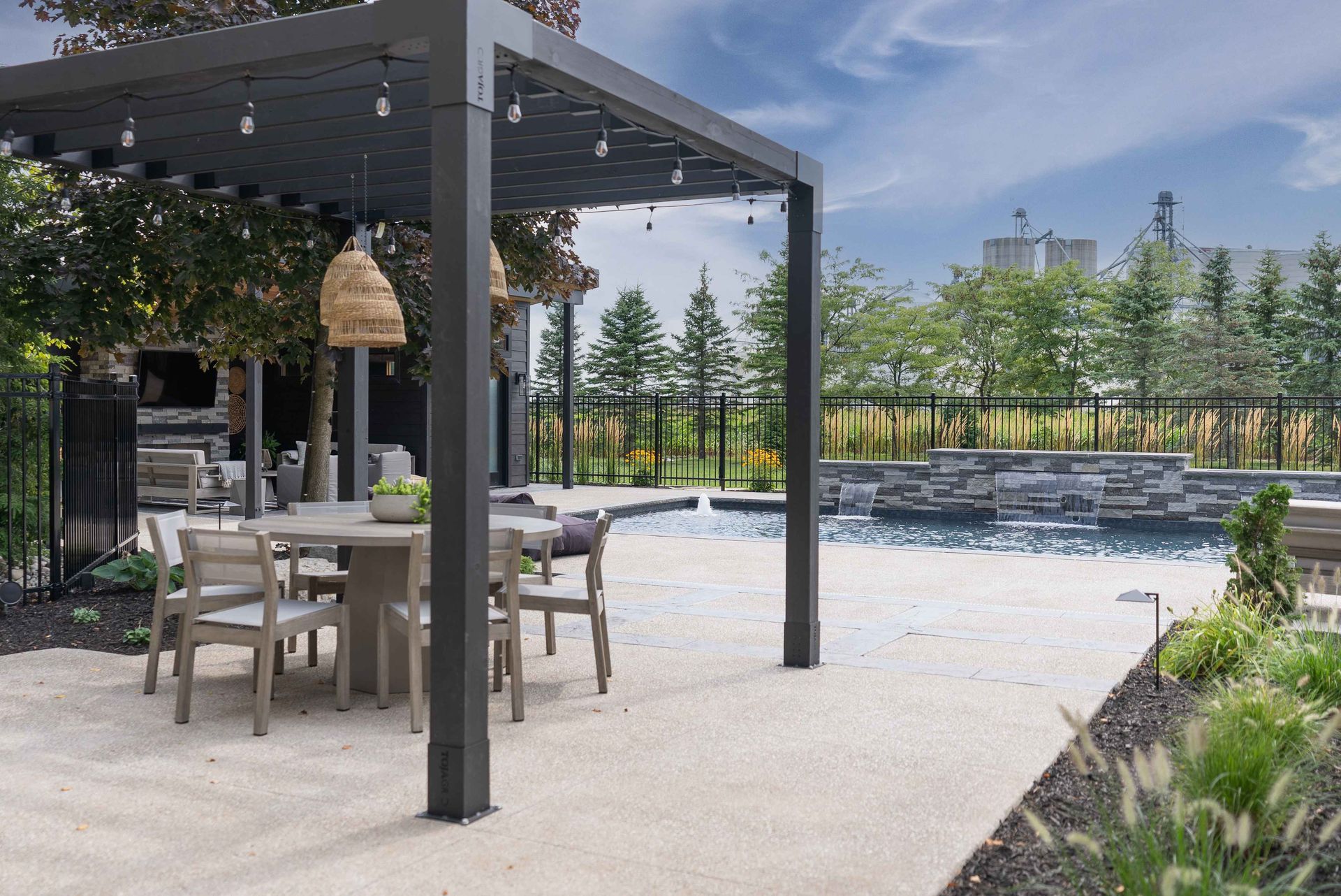 Outdoor patio with pergola, table and chairs beside a landscaped pool under a blue sky