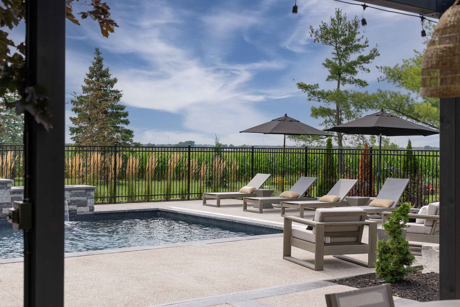 Poolside patio with lounge chairs, umbrellas, and a landscaped view beyond a black fence.