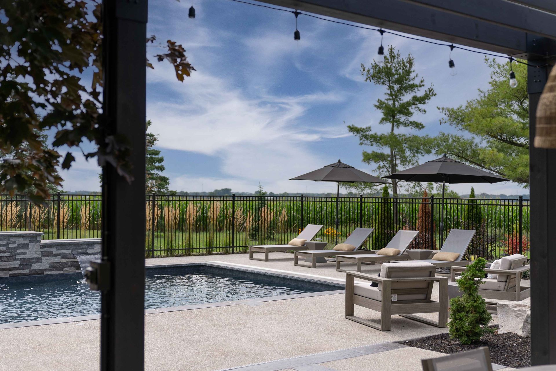 Covered patio with poolside lounge chairs, umbrellas, and a view of green fields under a blue sky