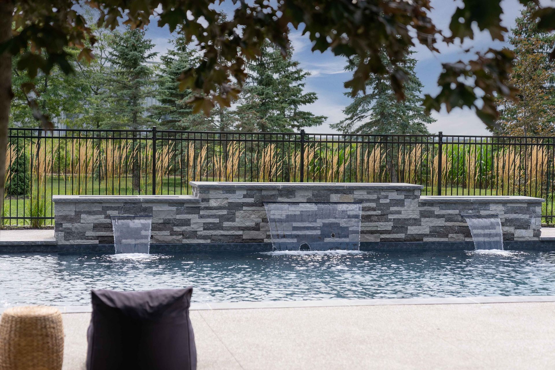 Backyard pool with stone water feature, black fence, trees, and a patio cushion in the foreground