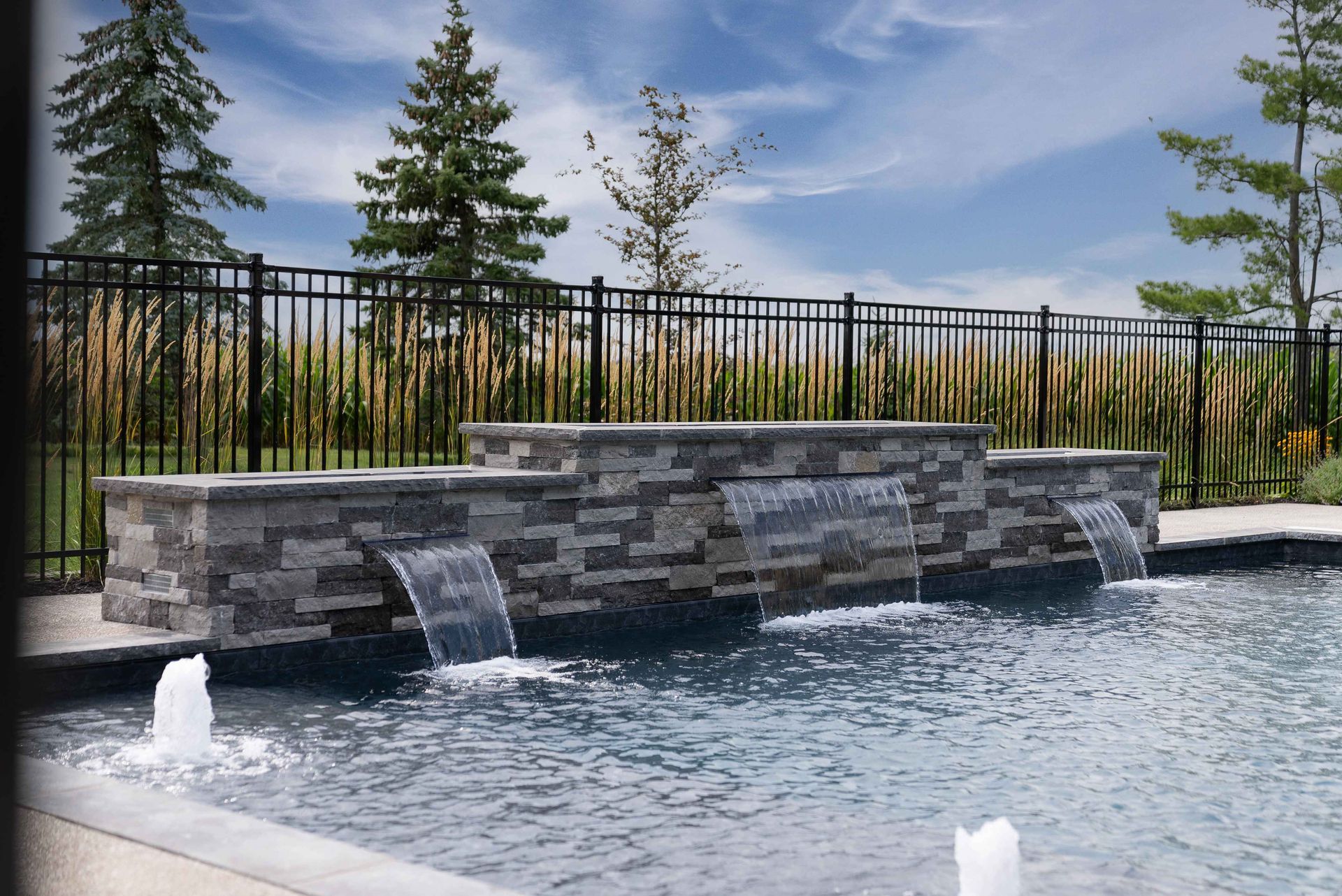 Stone waterfall feature in a landscaped pool with trees and a black fence behind it