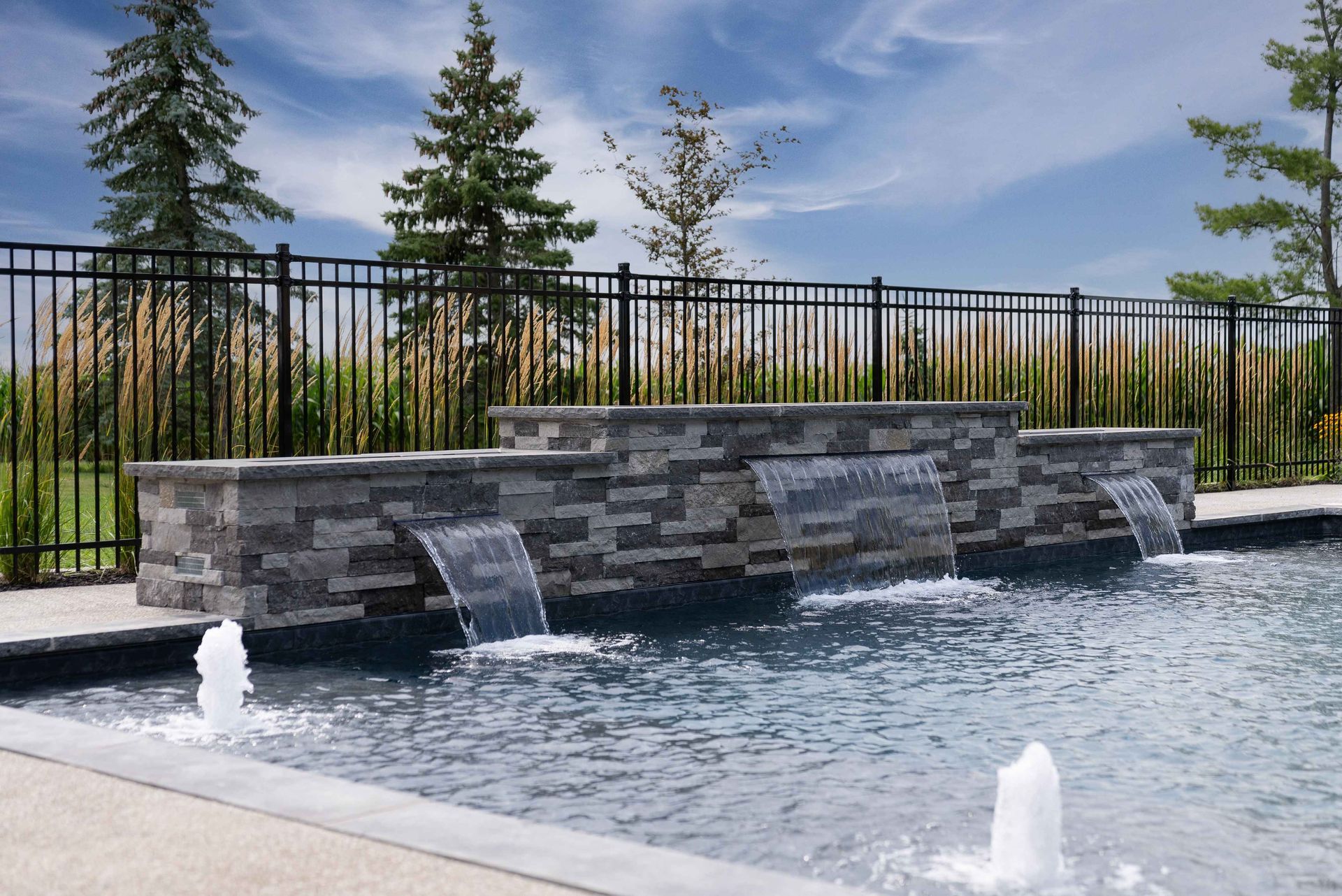 Stone fountain with water spilling into a pool, set beside a black fence and trees outdoors