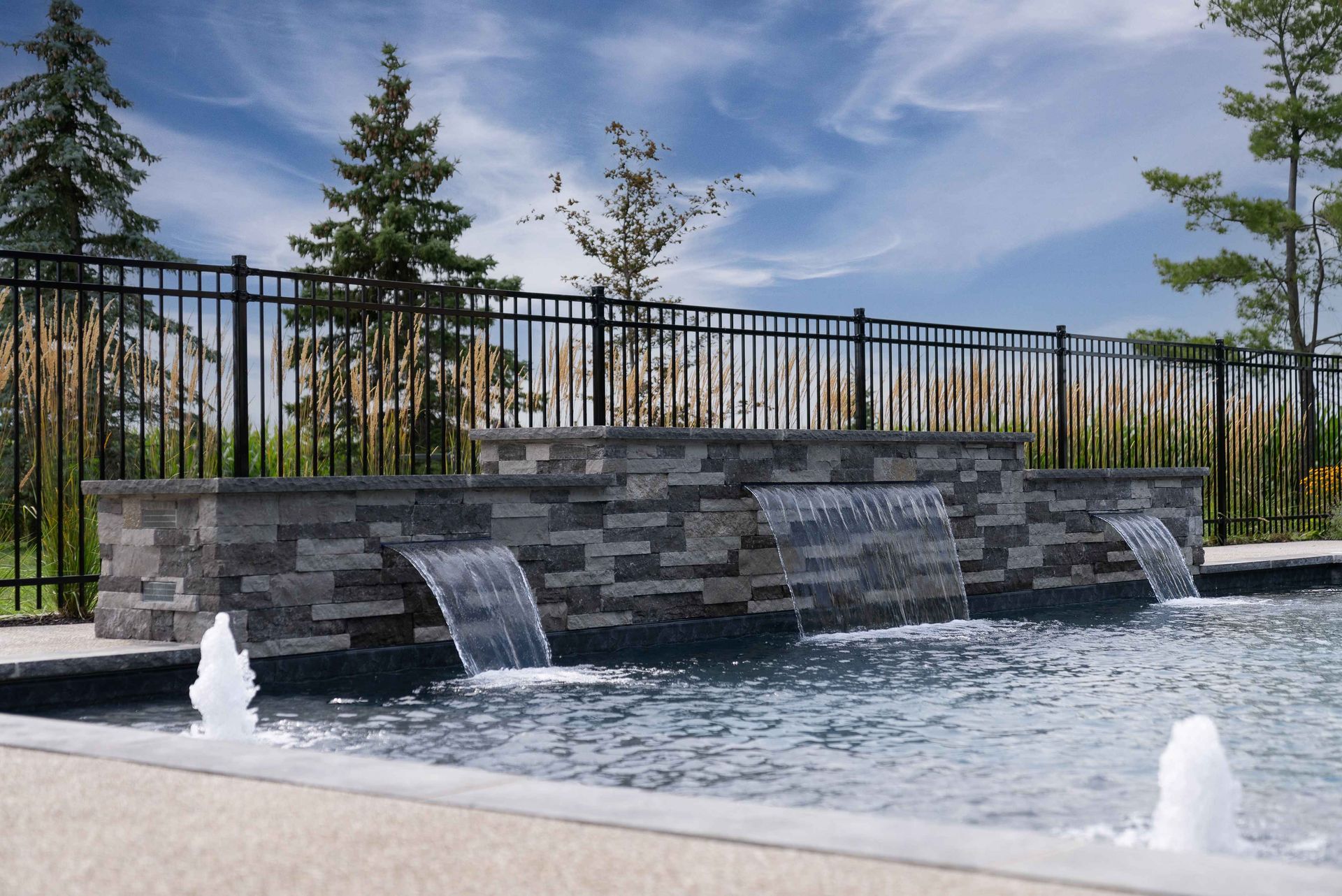 Modern stone water feature with cascading water and fountain jets by a fenced pool under a blue sky
