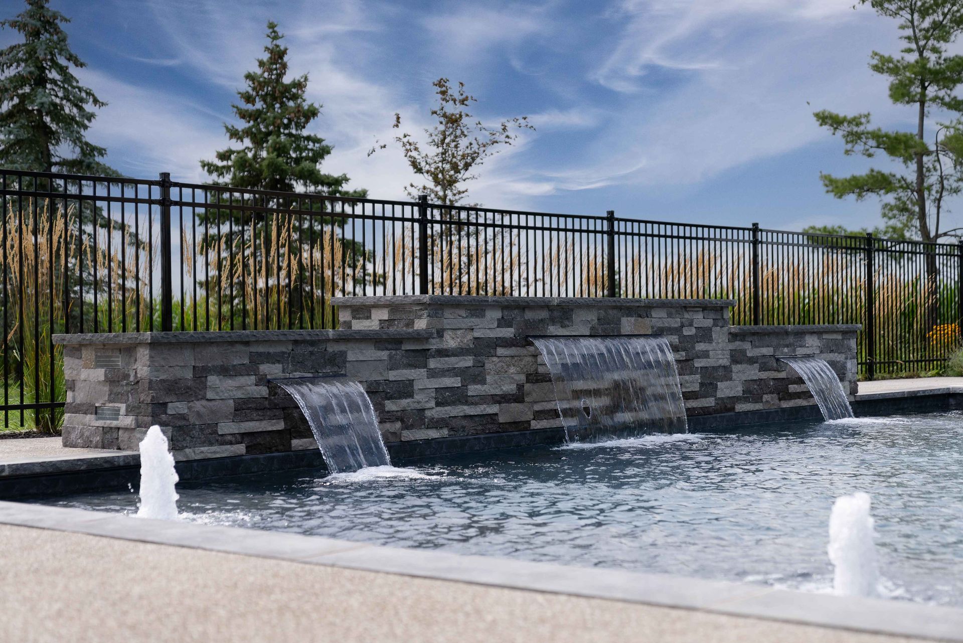 Modern outdoor fountain with stone wall, cascading water, and a black fence under a blue sky