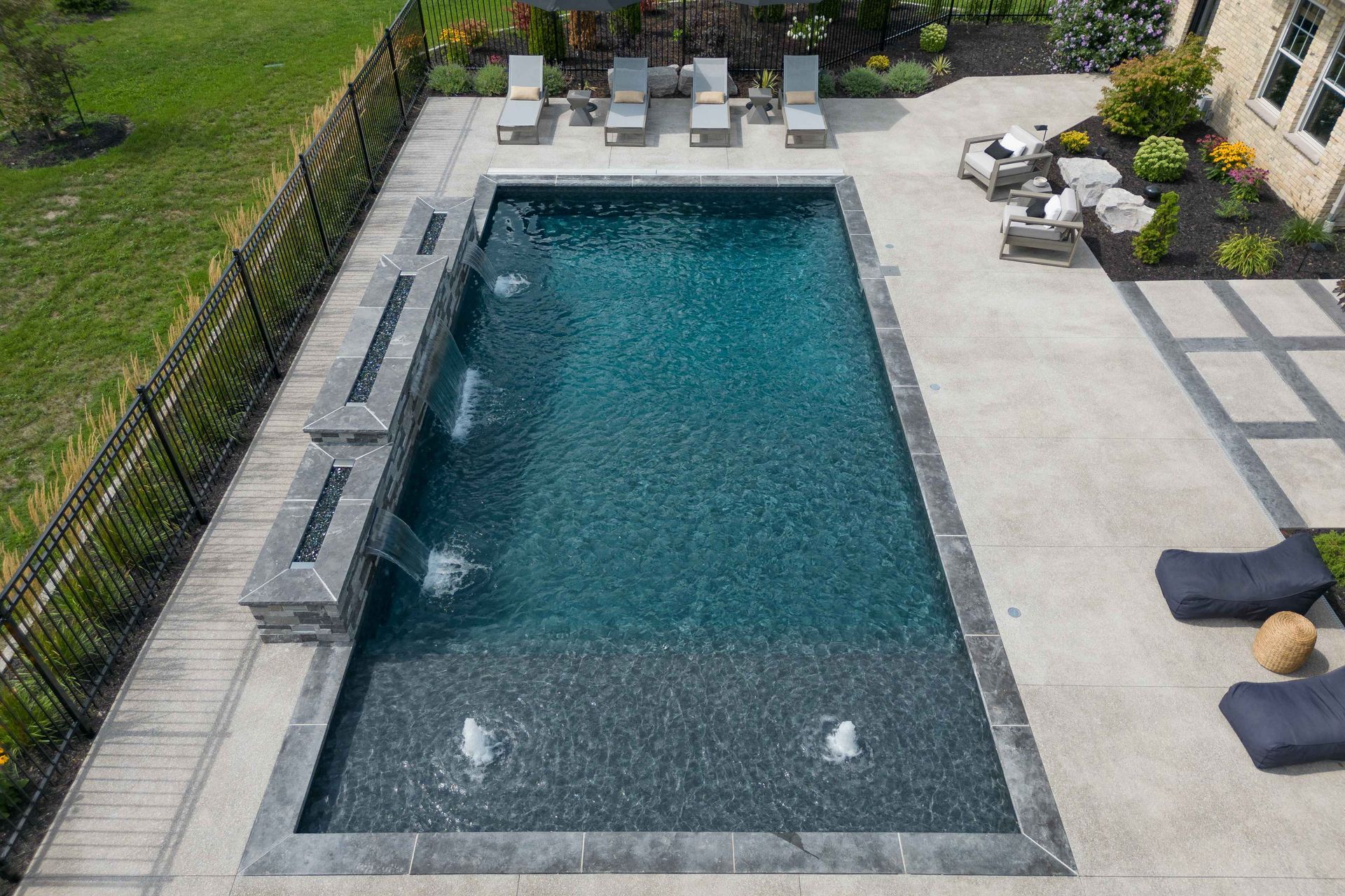 Rectangular backyard pool with lounge chairs and patio seating beside a black fence