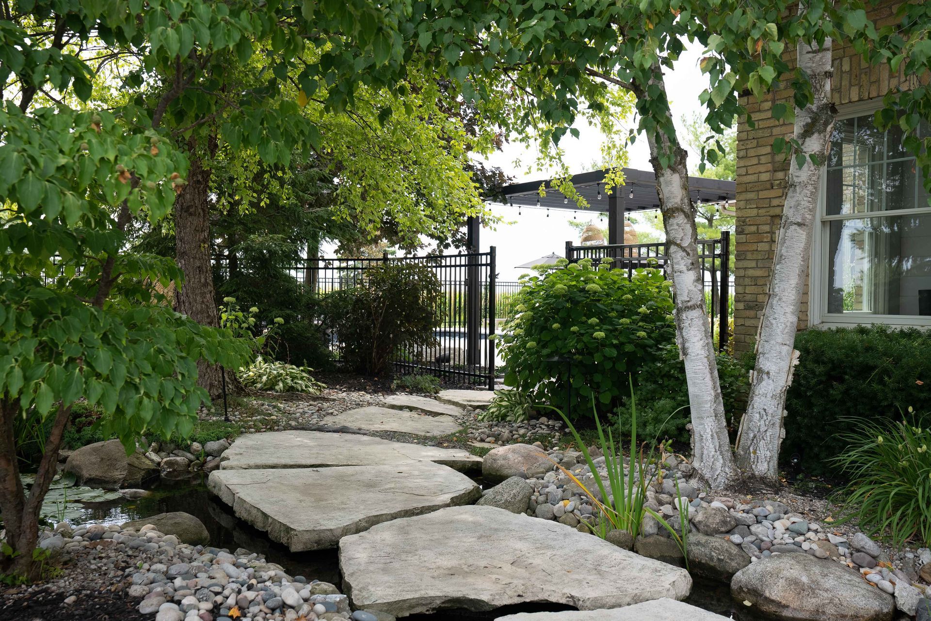 Stone path through a leafy garden leading to a black gate beside a house and trees
