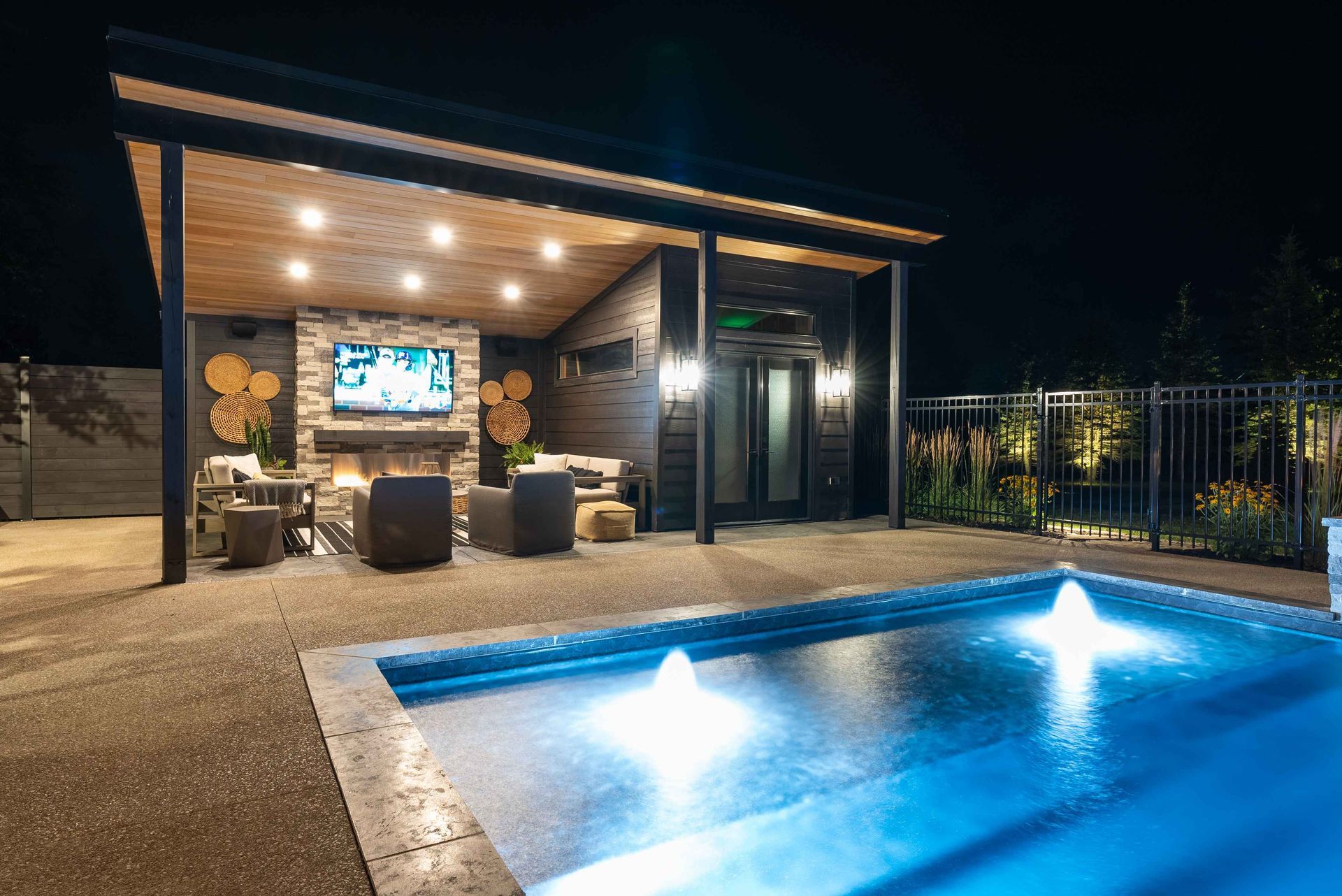 Modern backyard patio at night with lit pool, lounge seating, and a covered outdoor TV area