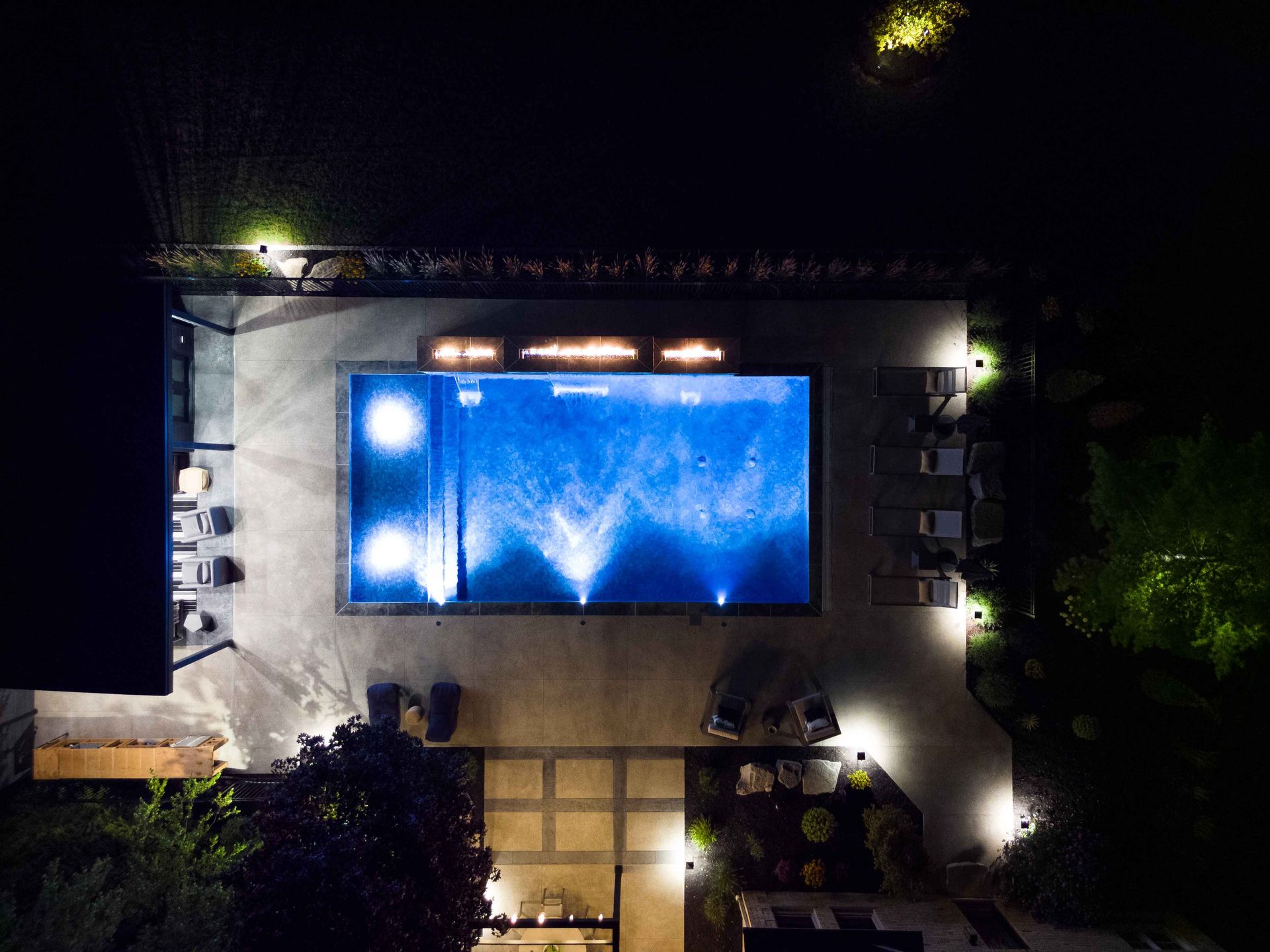 Nighttime aerial view of a lit rectangular swimming pool beside a modern house and dark garden.