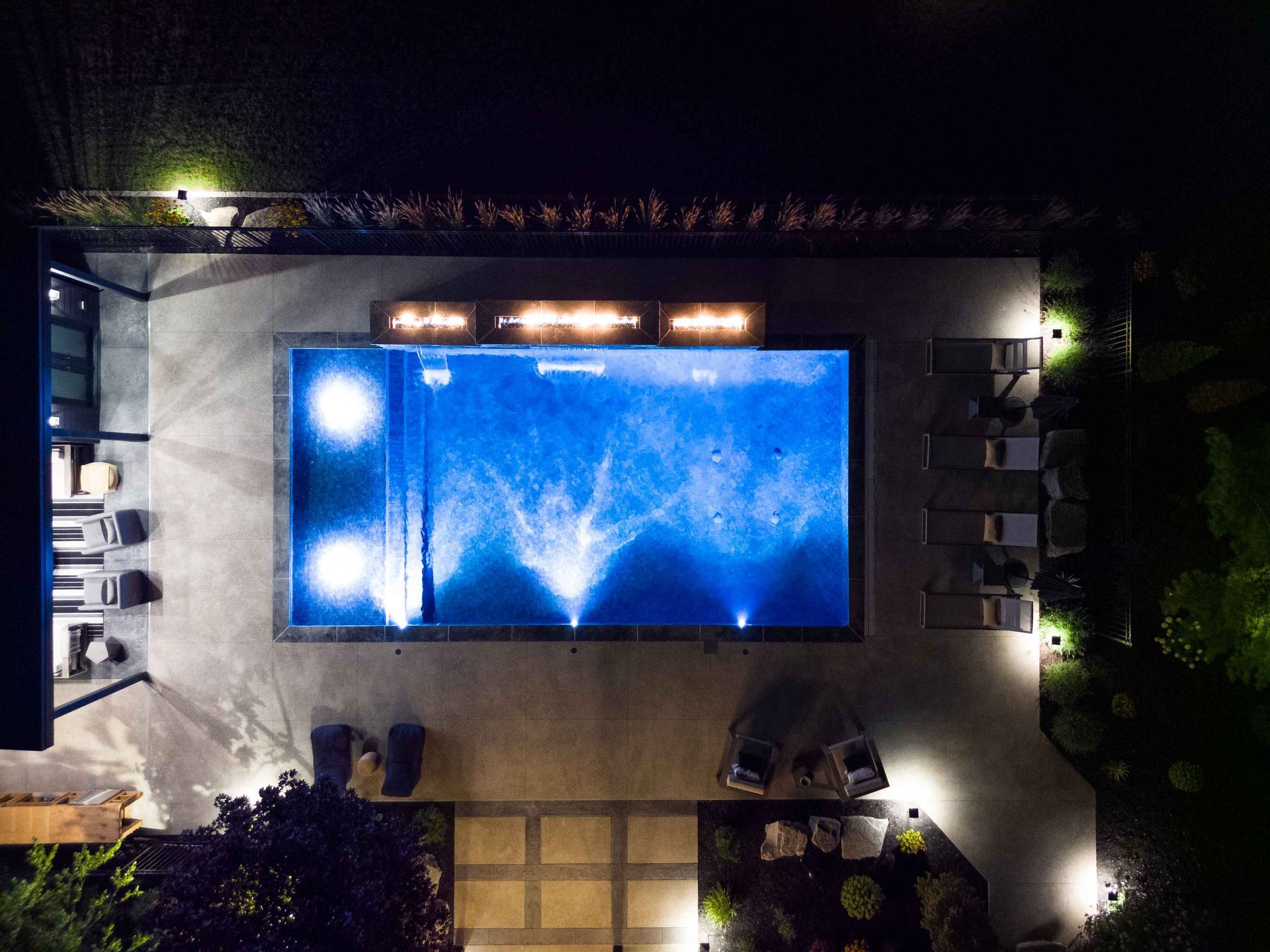Nighttime pool lit blue in a courtyard, surrounded by lounge chairs and trees.