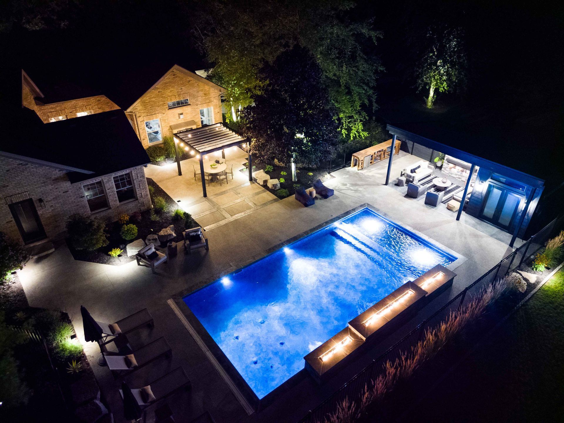 Nighttime aerial view of a lit backyard with a glowing blue pool, patio seating, and stone buildings
