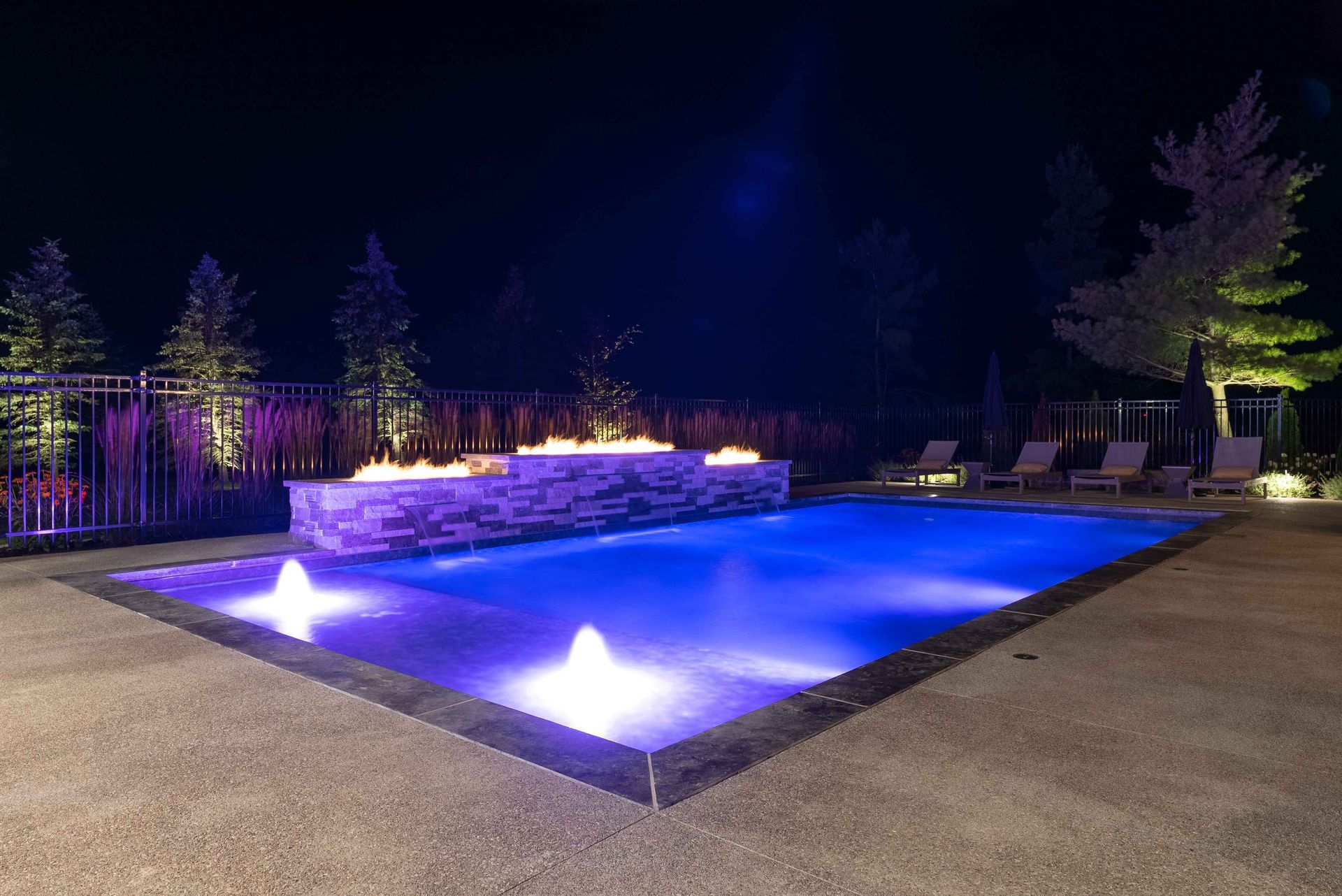 Illuminated backyard pool at night with purple and blue lights and a stone wall backdrop