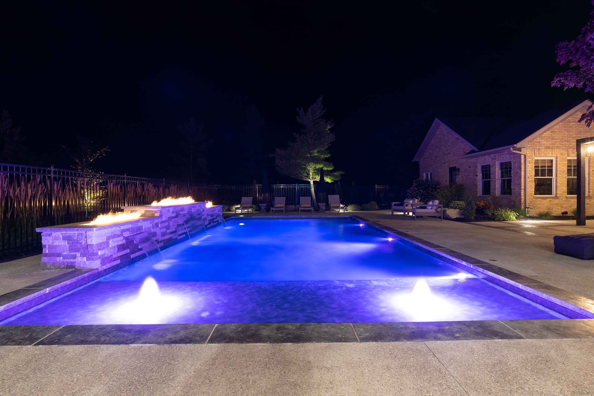 Blue-lit backyard swimming pool at night beside a house and stone patio with fountain jets on the water.