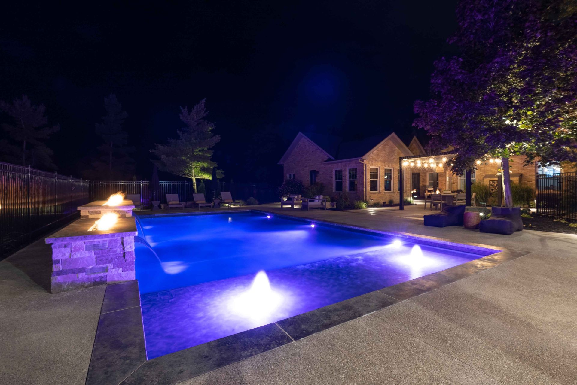 Blue-lit backyard swimming pool at night with patio lights and a house in the background