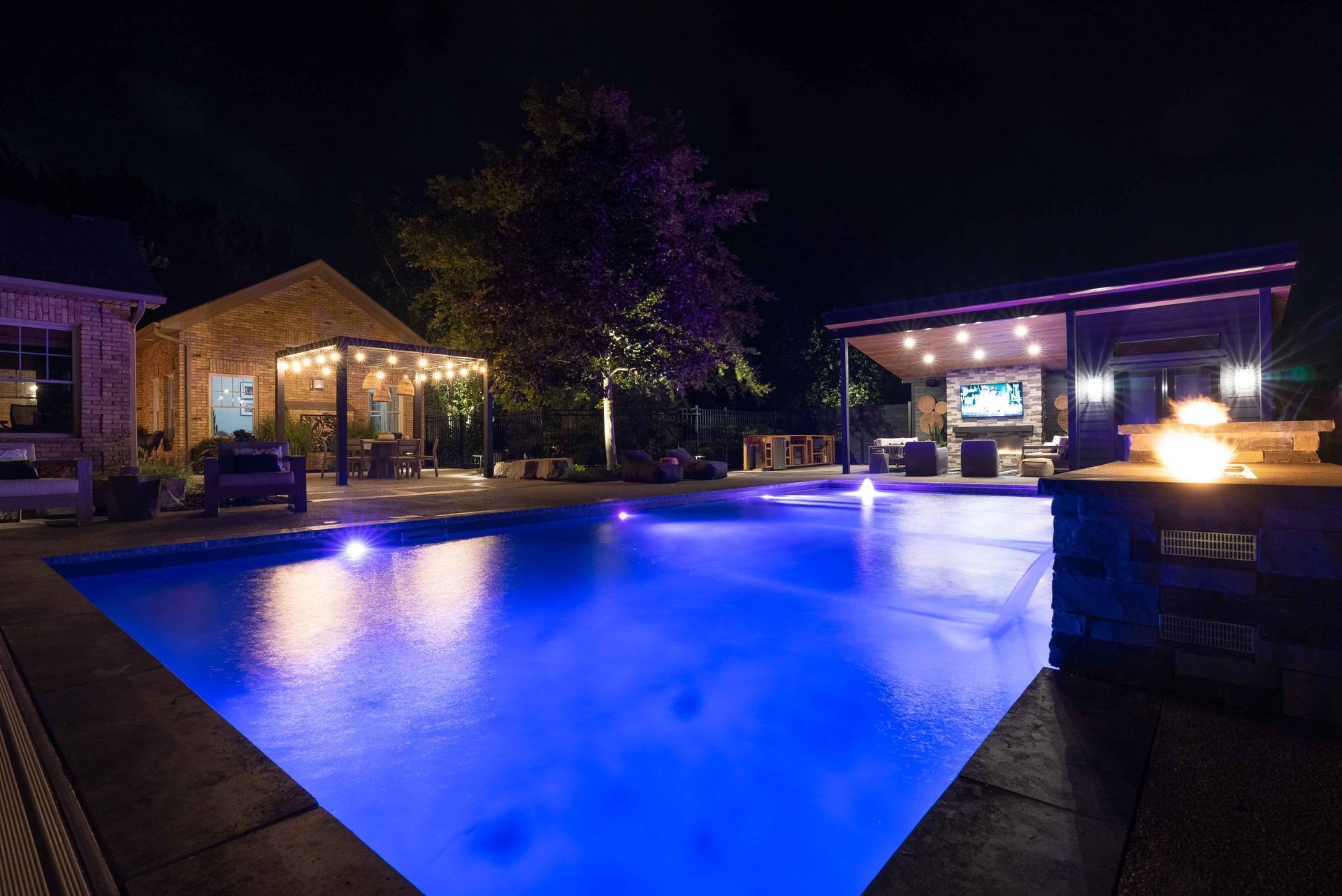 Illuminated outdoor pool at night with blue water, lounge chairs, and a lit patio area