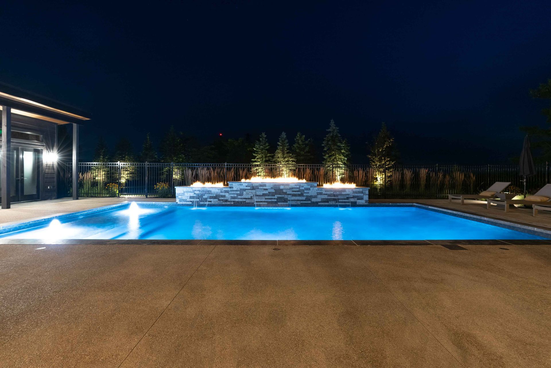 Illuminated backyard swimming pool at night with blue water, stone deck, and patio seating area