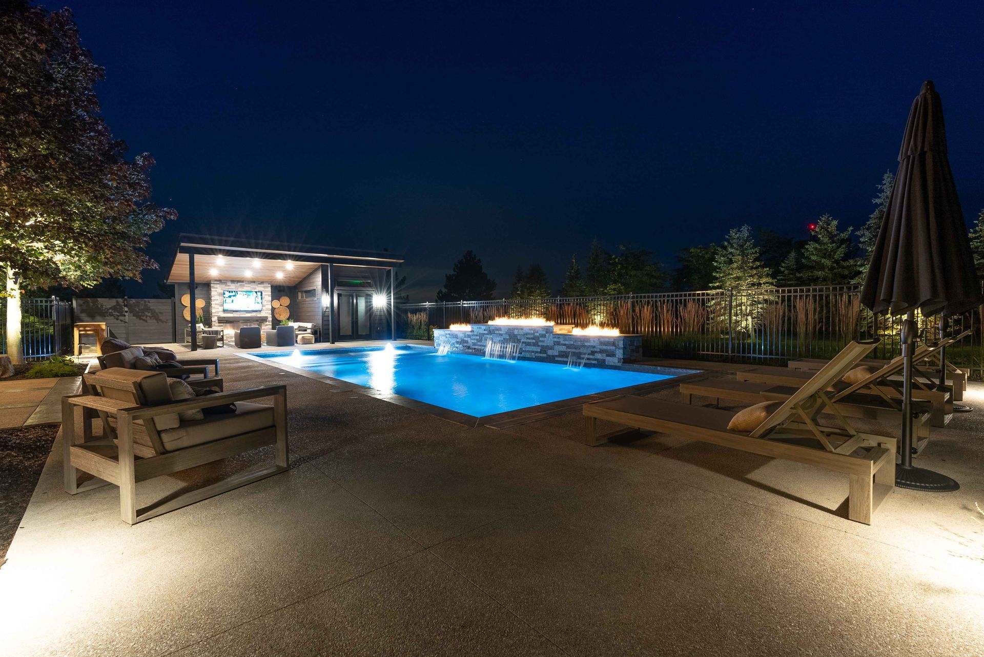 Illuminated backyard pool at night with lounge chairs and modern patio seating