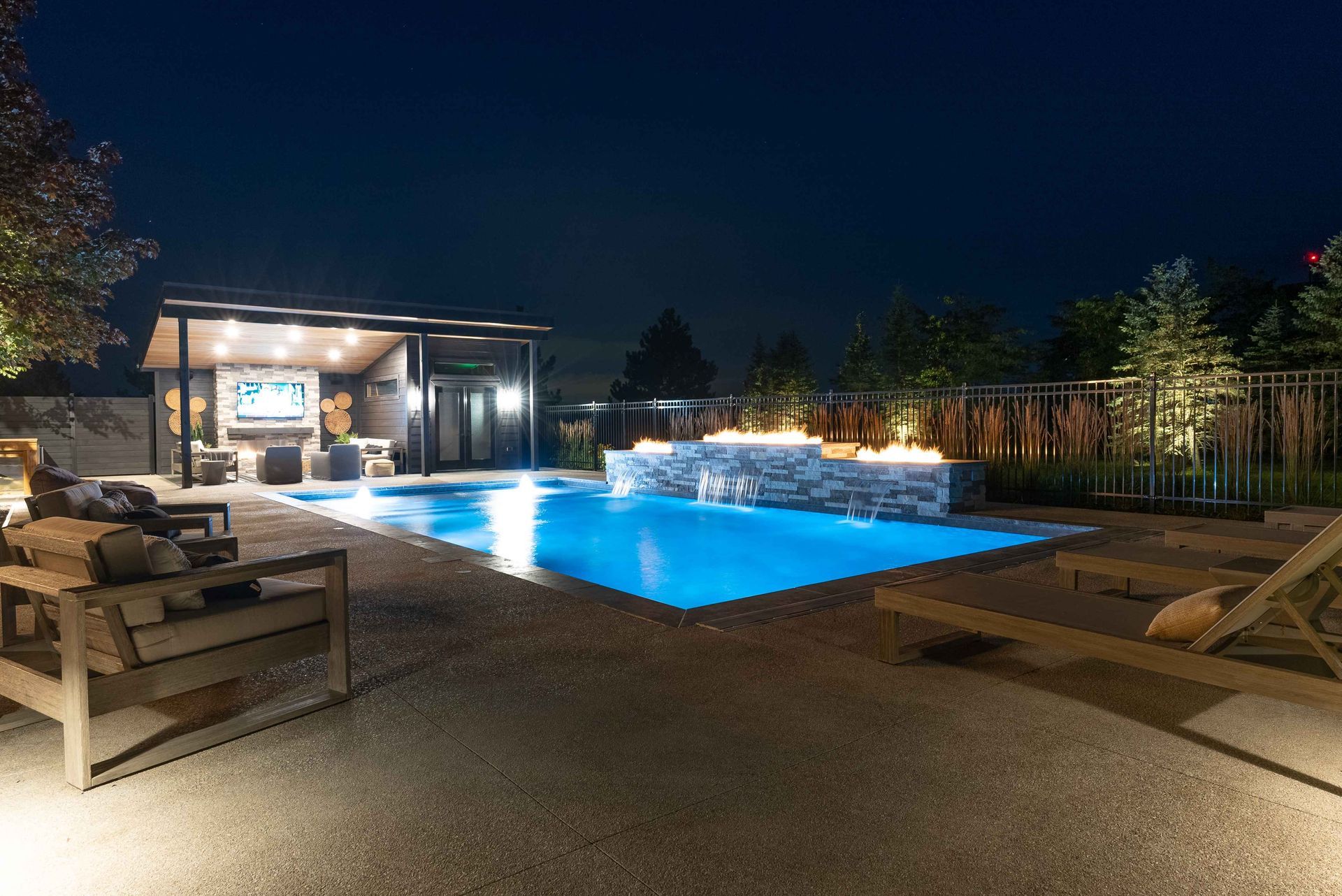 Nighttime backyard pool with glowing blue water, lounge chairs, and a lit patio seating area