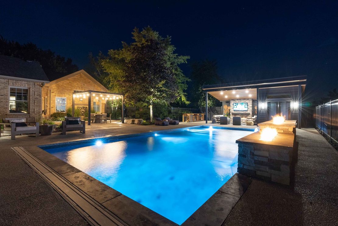 Illuminated outdoor pool beside a lit building at night, with blue water and lounge area