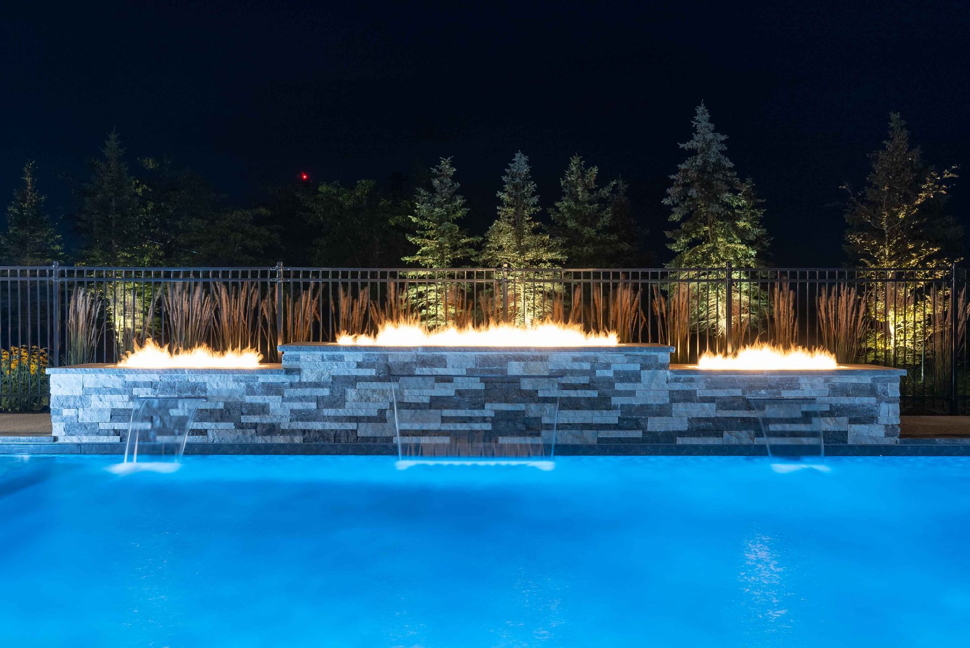 Illuminated backyard pool with stone waterfall, fire features, and lit trees at night