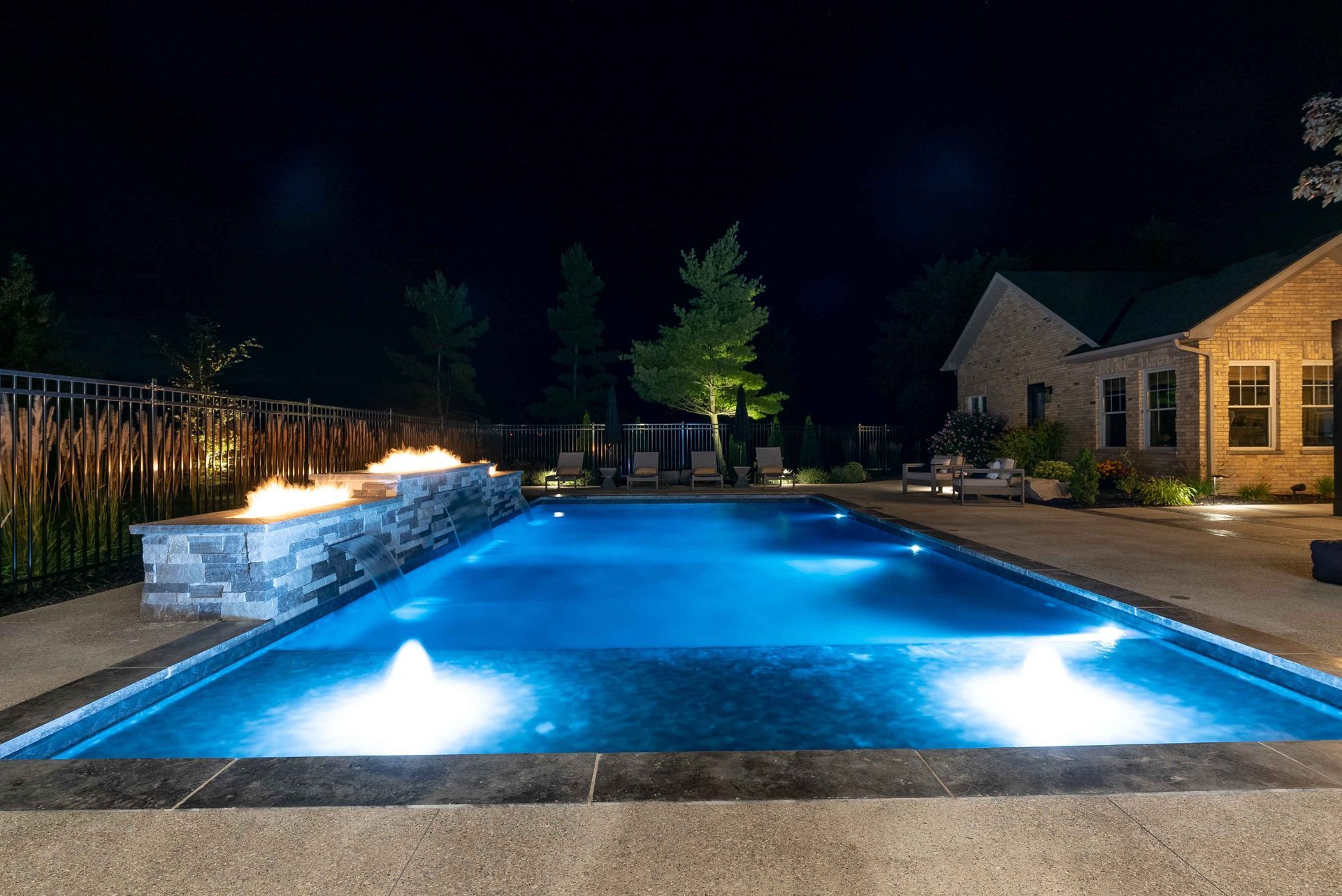 Nighttime backyard with a lit blue swimming pool beside a house and patio seating